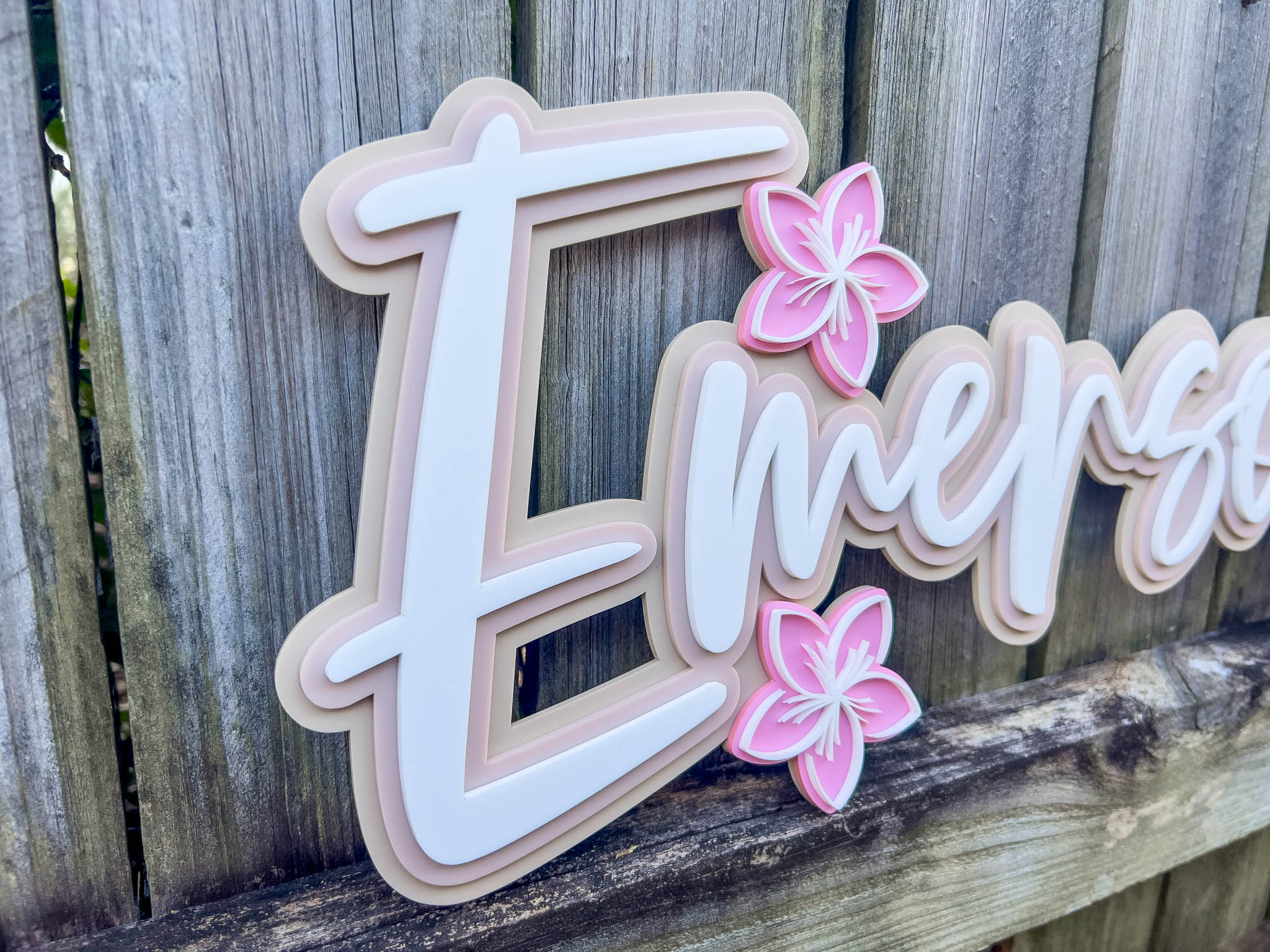 Close-up of a 3D name plaque with 'Emerson' and pink frangipanis on a wooden background