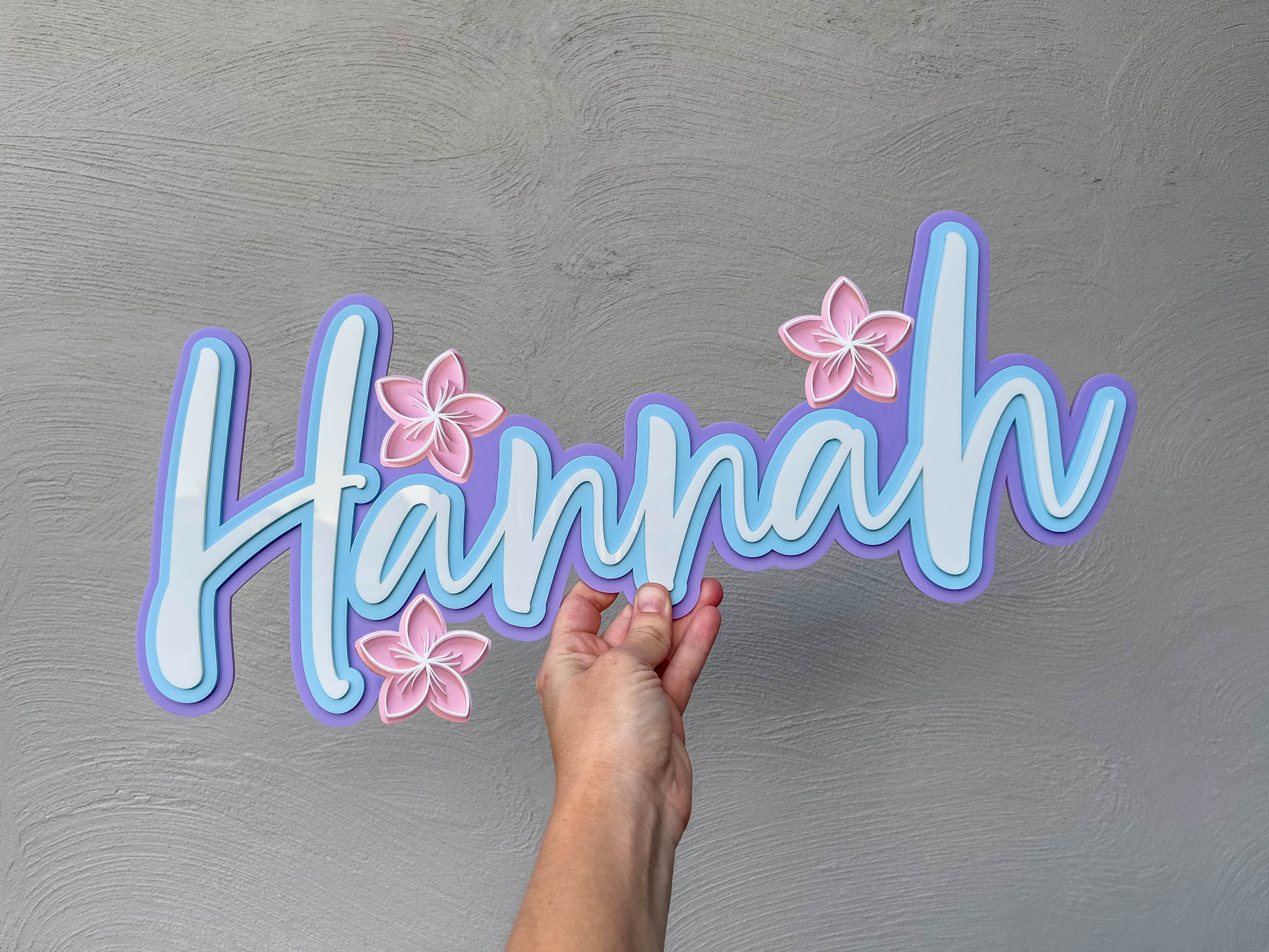 Elegant handwritten style custom name sign with 'Hannah' in light blue, purple and white with light pink frangipanis.