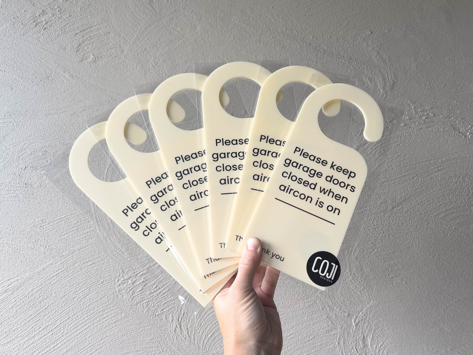 Hand holding multiple 'Please keep garage doors closed when aircon is on' signs against a grey background | Coji Designs