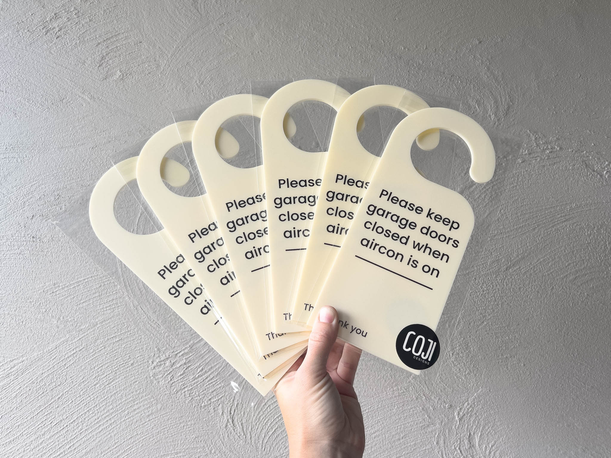 Hand holding multiple 'Please keep garage doors closed when aircon is on' signs against a grey background | Coji Designs