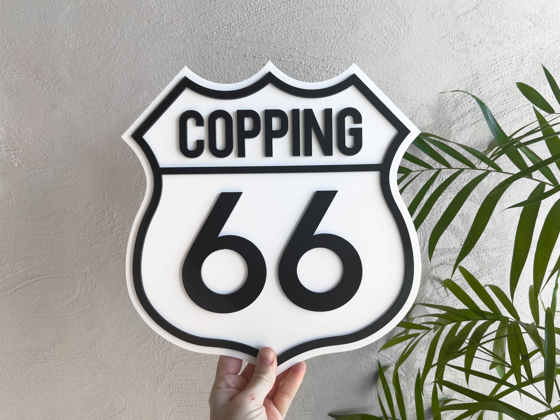 Decorative badge style sign with 'Copping 66' text held against a neutral wall with plants.