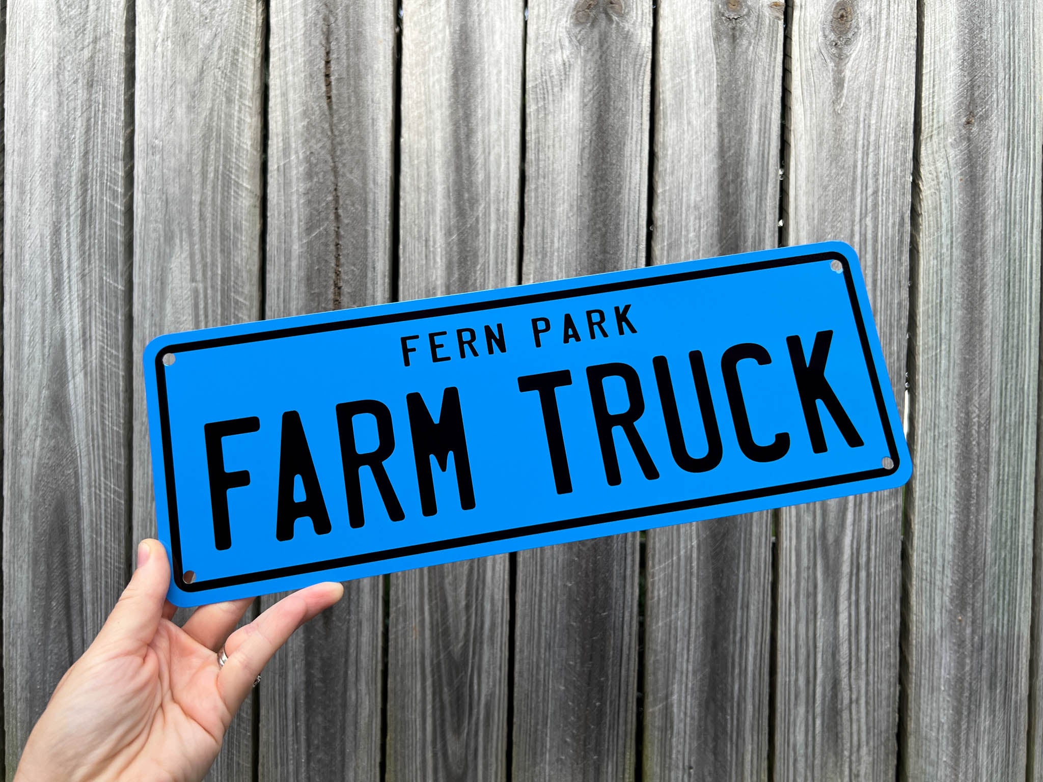 Hand holding a blue number plate sign with 'Fern Park Farm Truck' text against a wooden background.