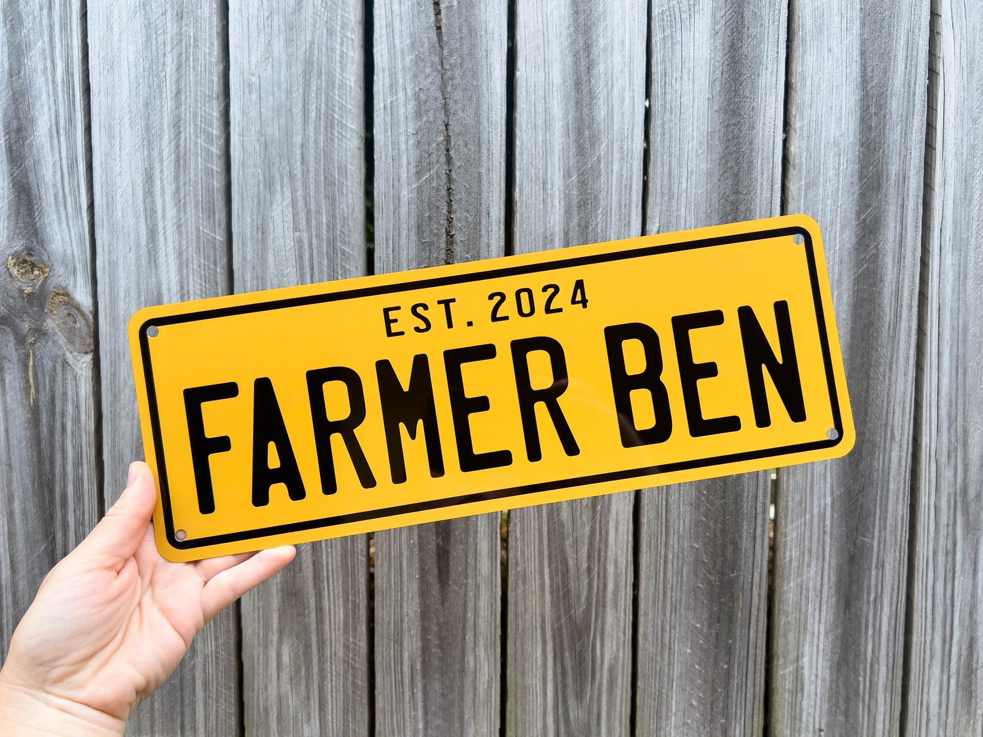 Yellow aluminium sign with 'FARMER BEN' and 'EST. 2024' text held against a wooden fence background