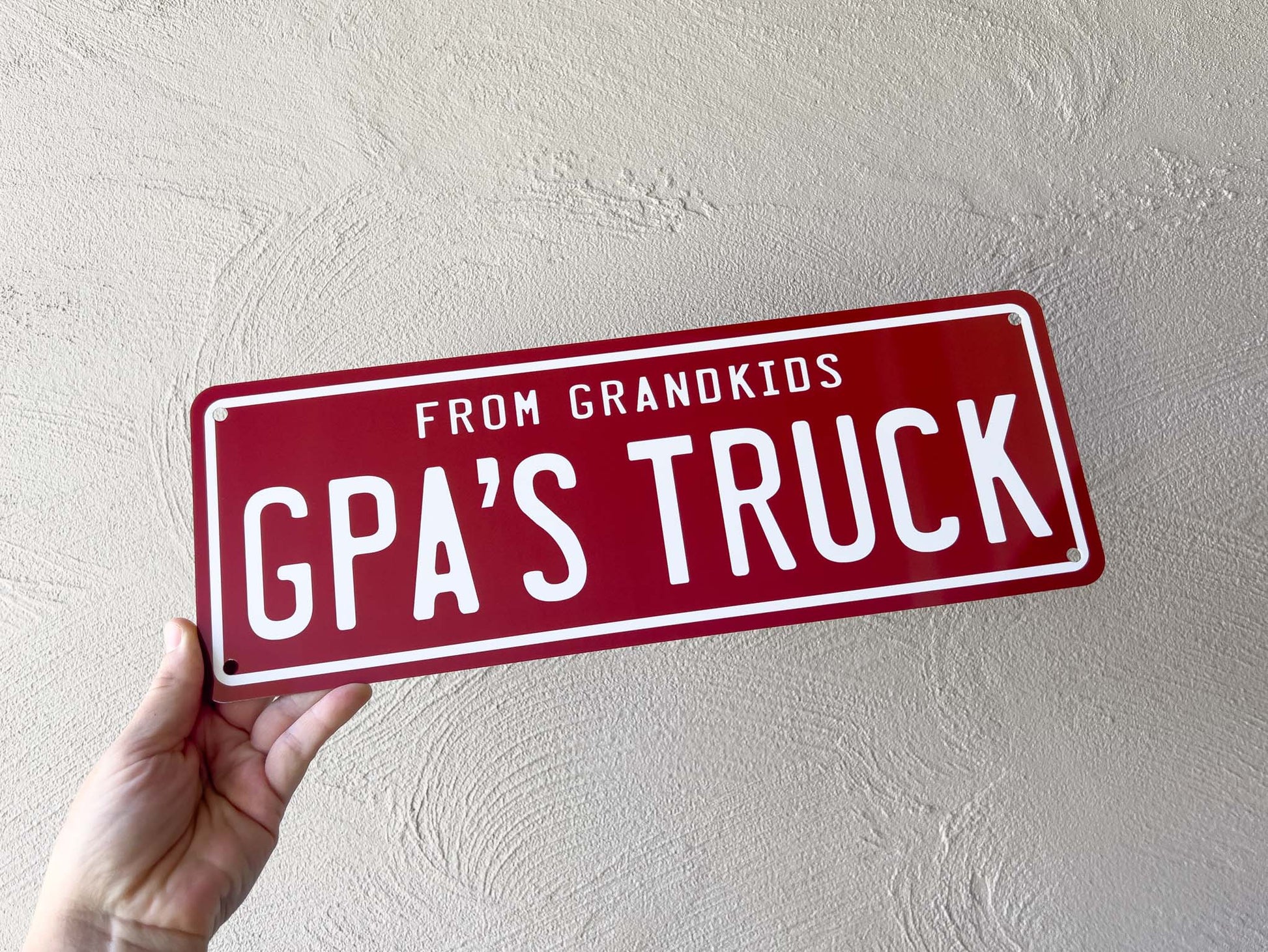 Hand holding a maroon sign with white text 'FROM GRANDKIDS GPA'S TRUCK' against a light grey background