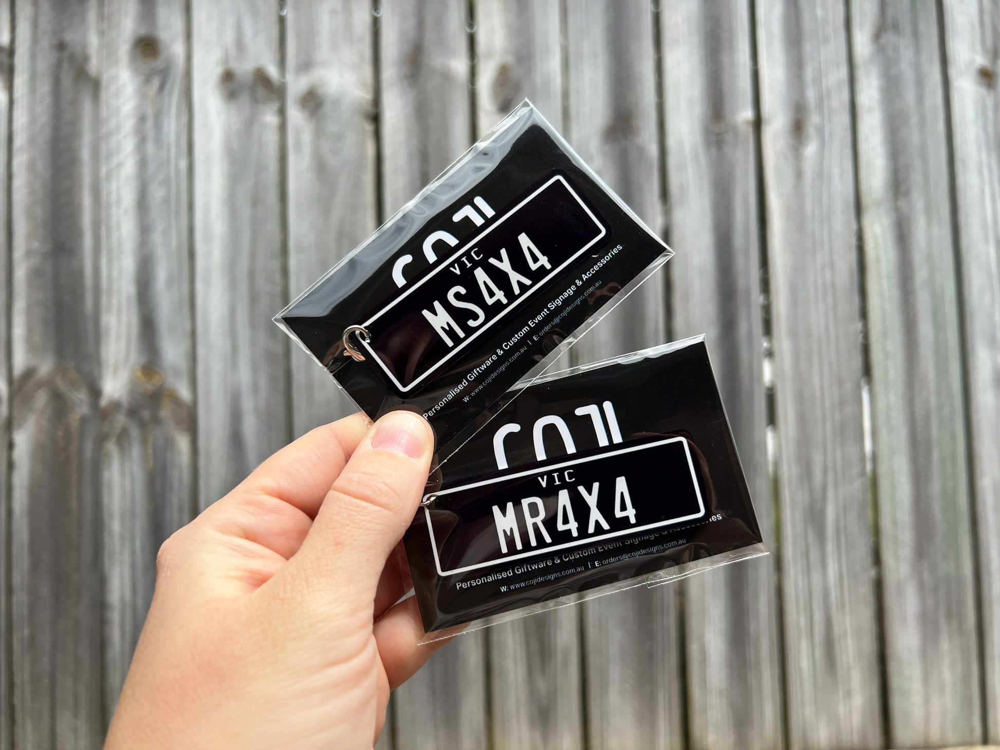 Hand holding two black license plate-style keychains with text against a wooden fence background