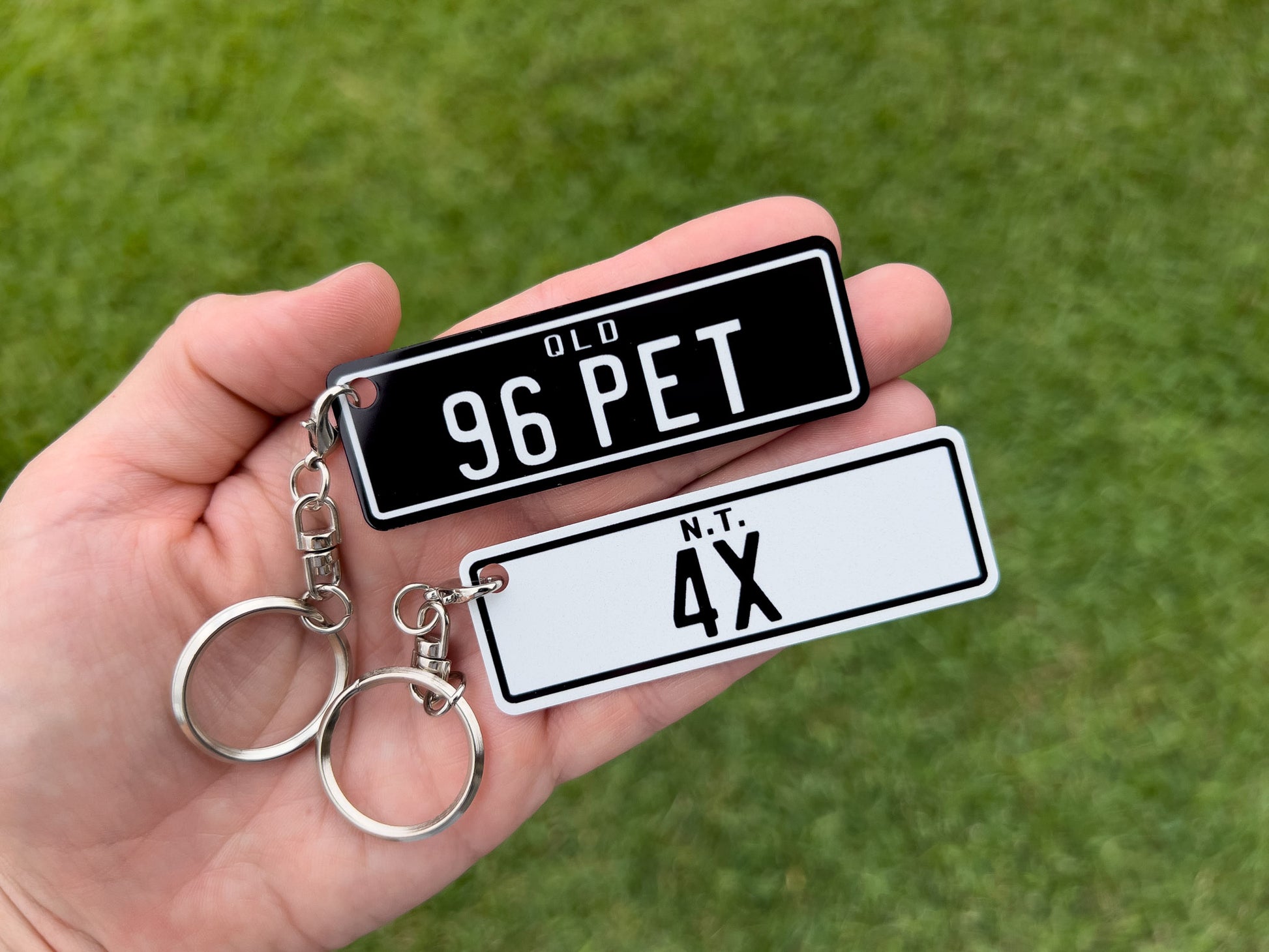 Hand holding three keychains with license plate designs against a grass background