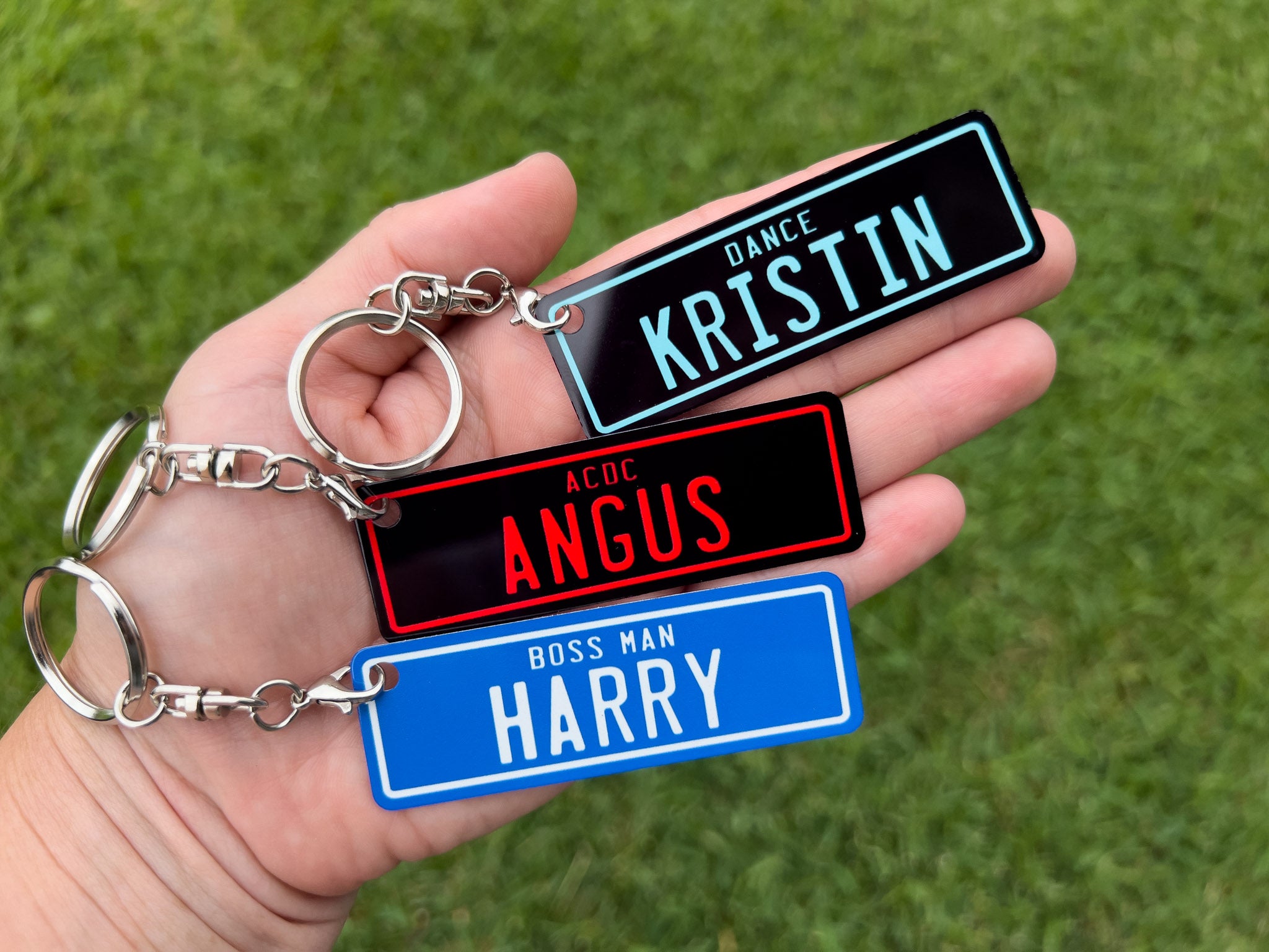 Hand holding three personalized keychains with names on a grass background