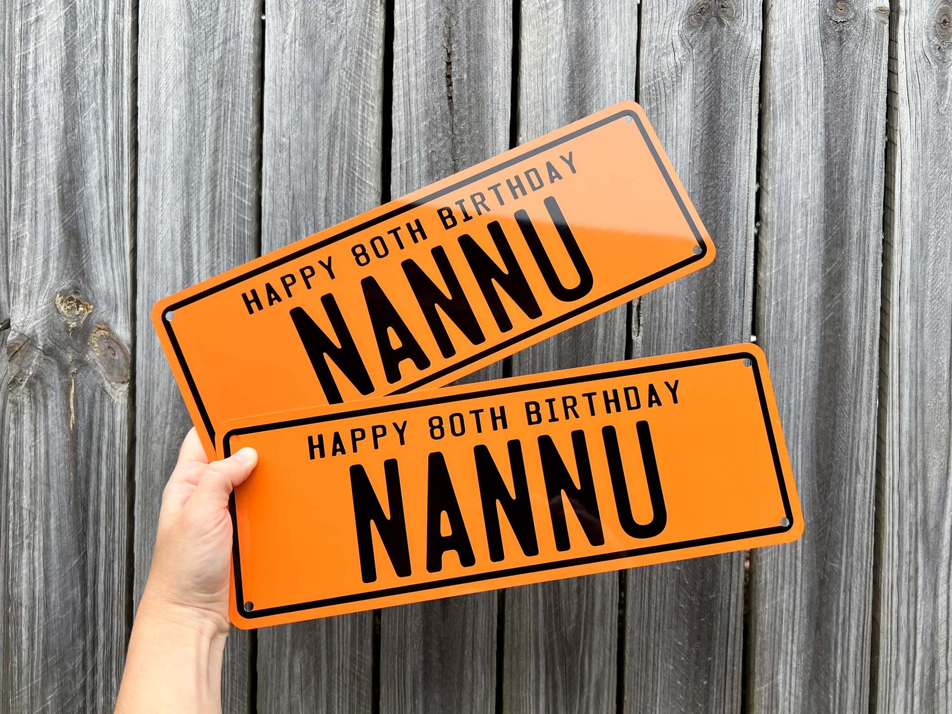 Two orange number plate signs with 'Happy 80th Birthday Nannu' text held against a wooden background.