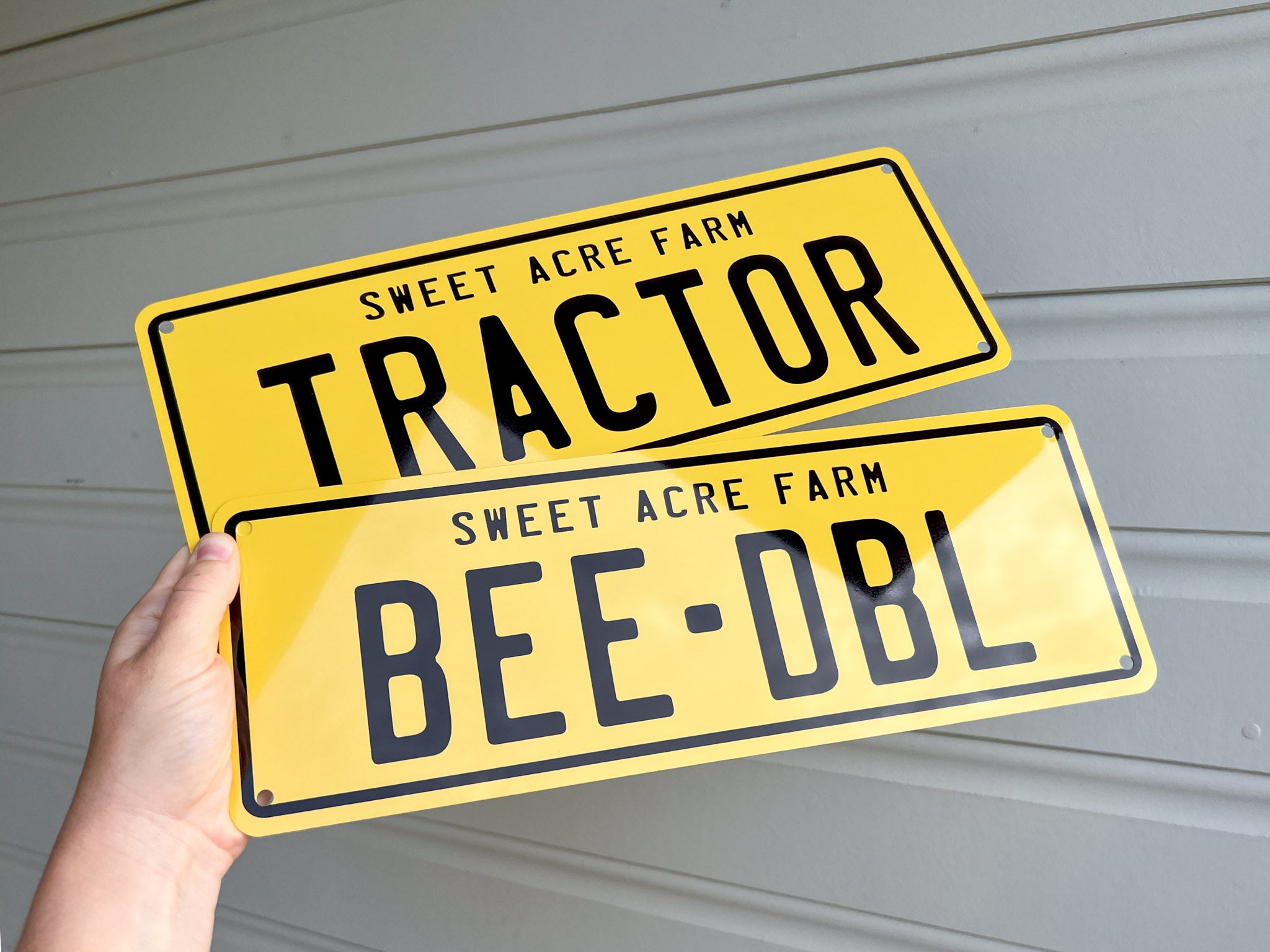 Two yellow metal signs with black text that say 'TRACTOR' & 'BEE-DBL' held by a hand against a grey background.