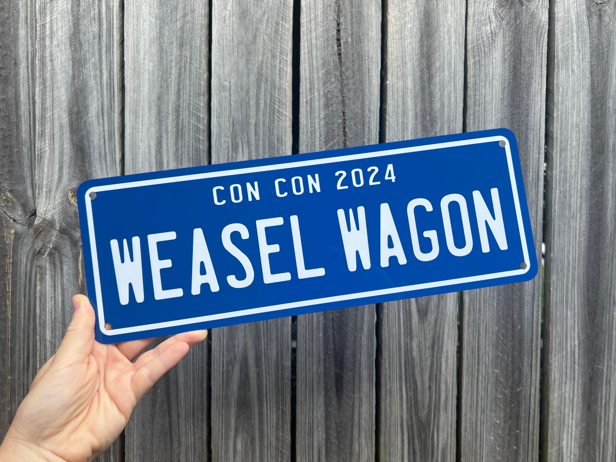 Coastal blue and white novelty number plate sign with 'CON CON 2024 WEASEL WAGON' text held against a wooden fence background