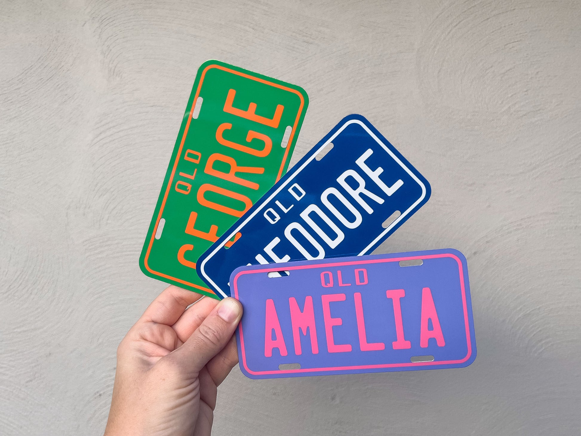 Three colorful license plate-shaped signs with names held by a hand against a grey background