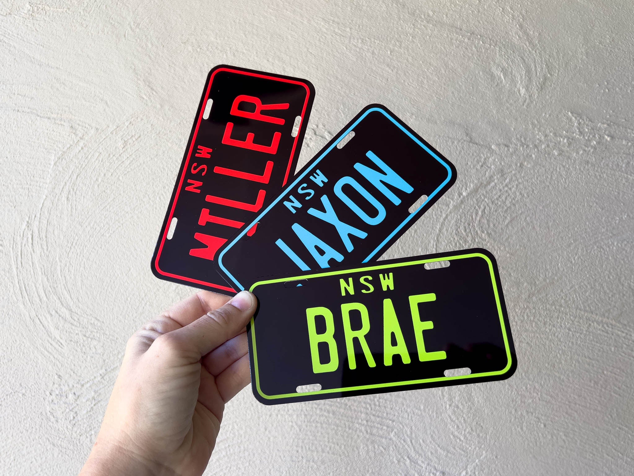 Three novelty mini number plates with text held by a hand against a light grey background