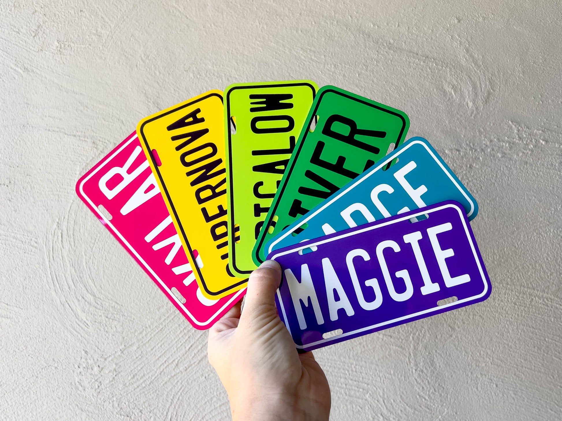 Hand holding a collection of colorful license plate-style name tags against a light grey background