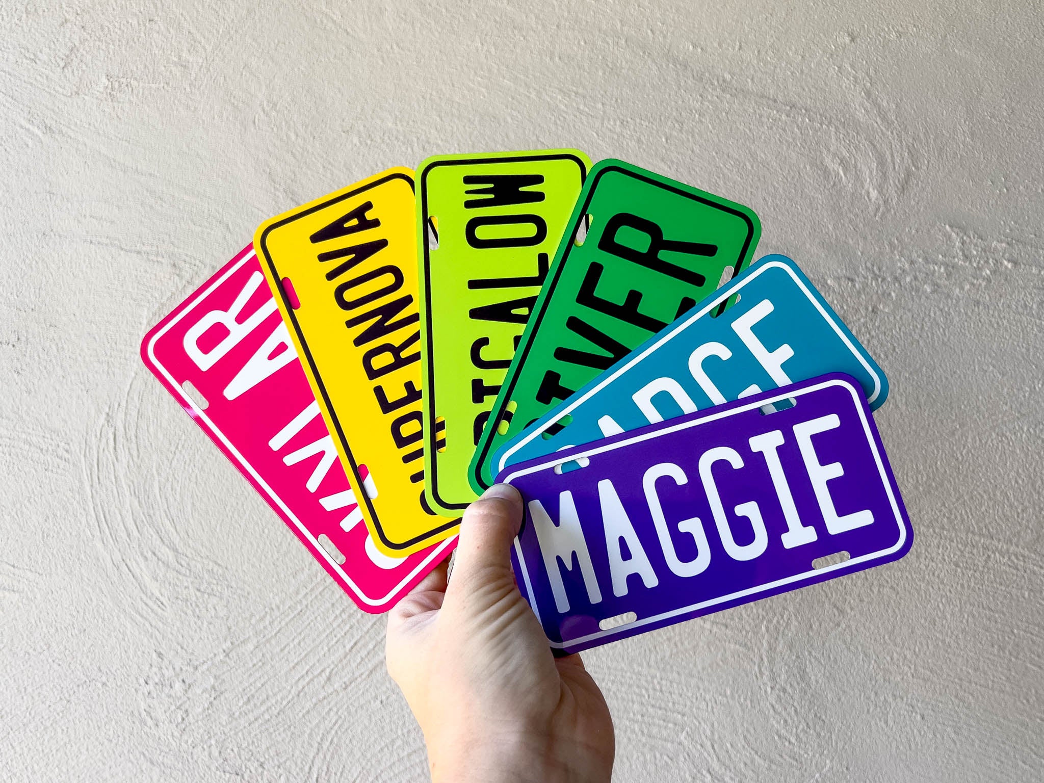 Hand holding a collection of colorful license plate-style name tags against a light grey background