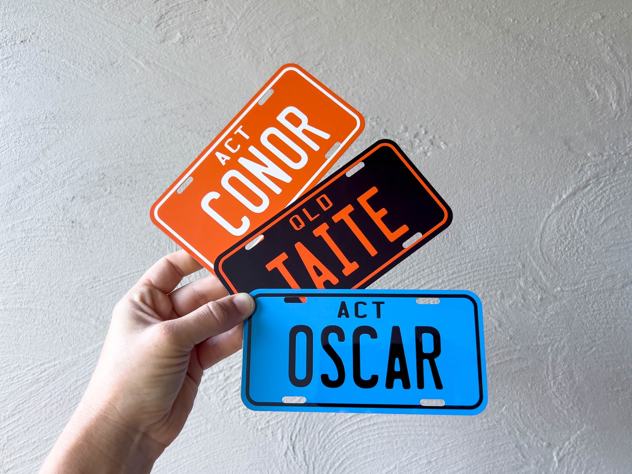Three mini license plate-shaped signs with names 'Conor', 'Taite', and 'Oscar' held by a hand against a light grey background.
