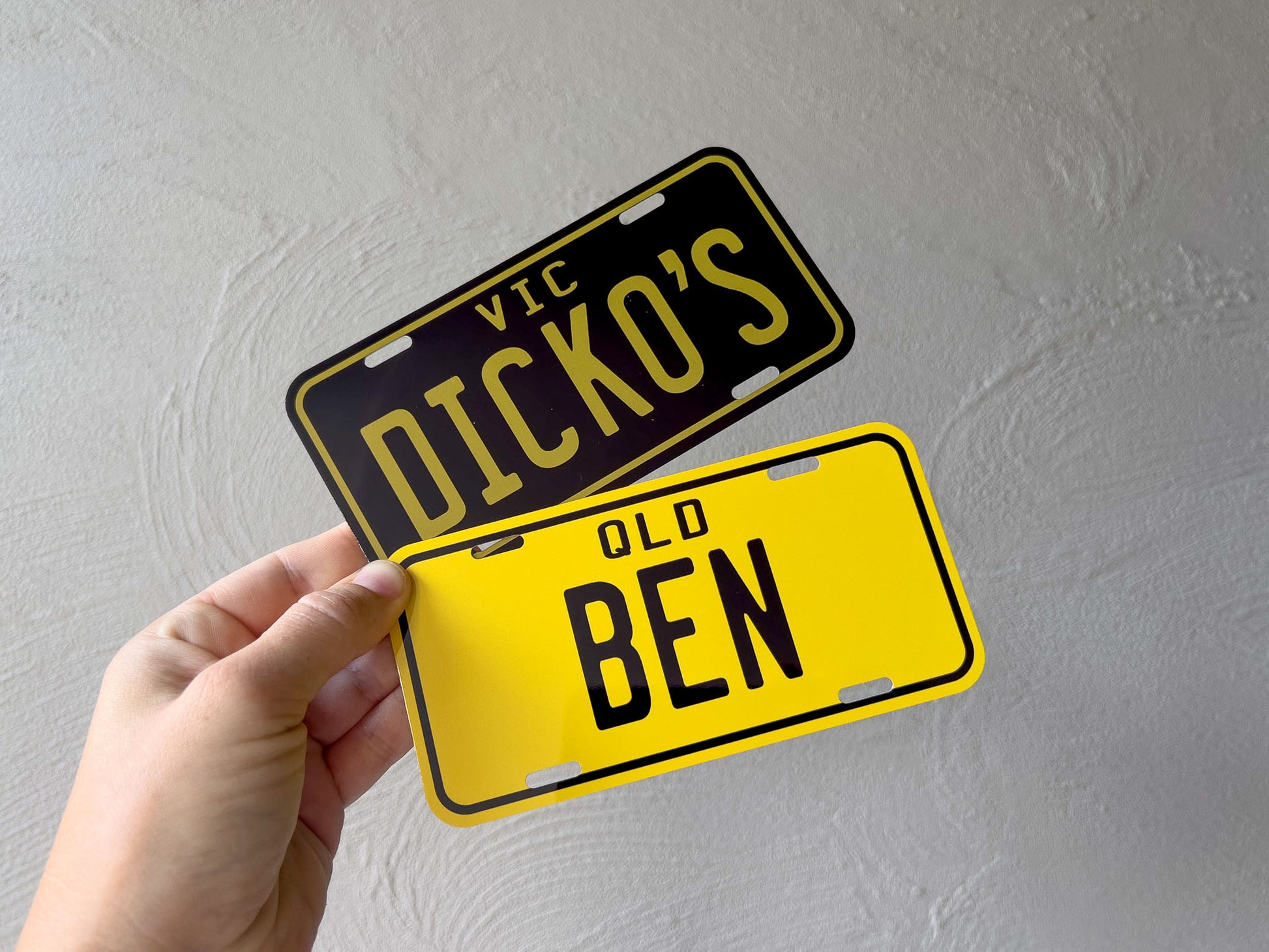 Two mini novelty license plates in yellow and black held against a plain background