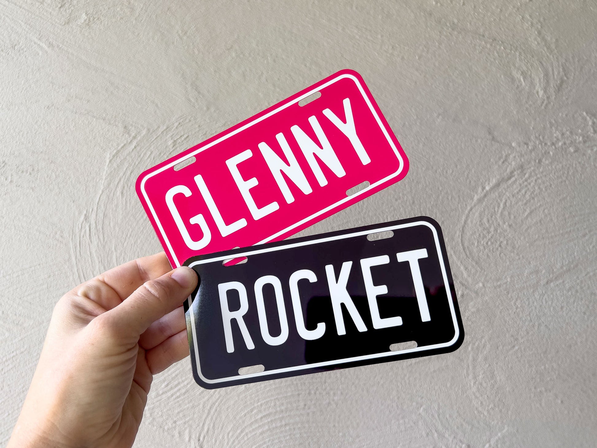 Two license plates, one pink with 'GLENNY' and one black with 'ROCKET', held against a light grey background.