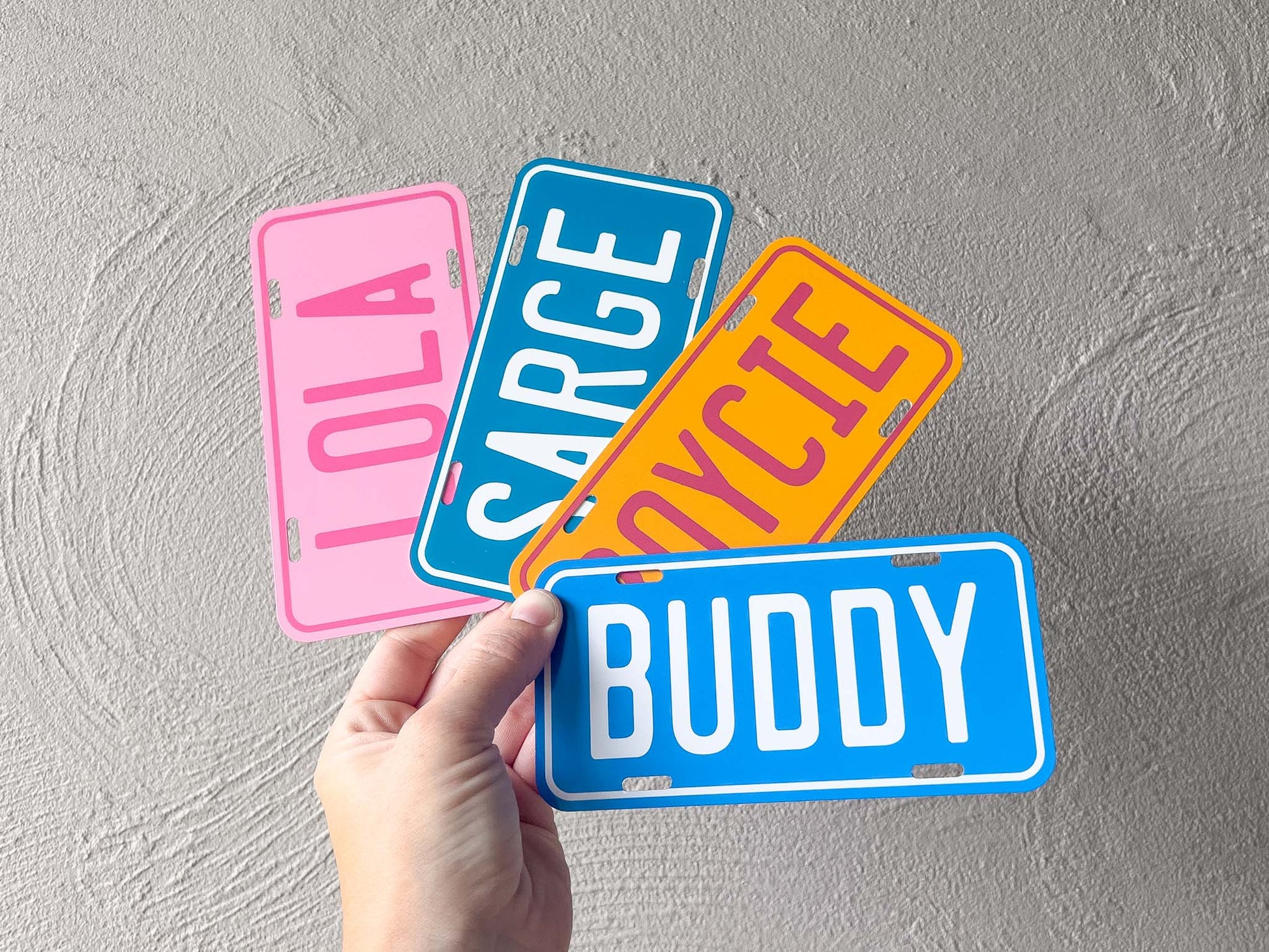 Four colorful license plate-style name tags held by a hand against a grey background.