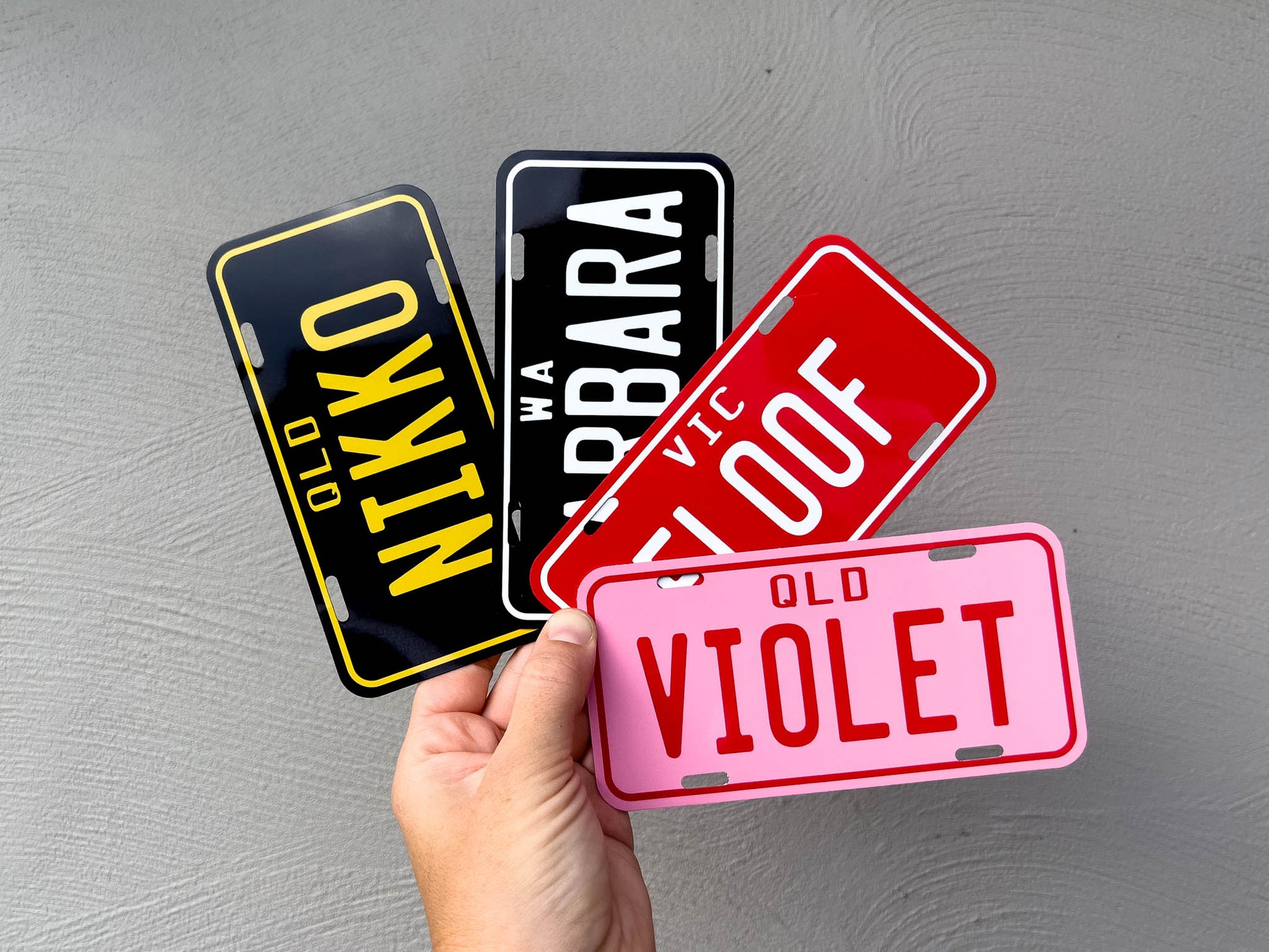 Four license plate-style name signs held by a hand against a grey background