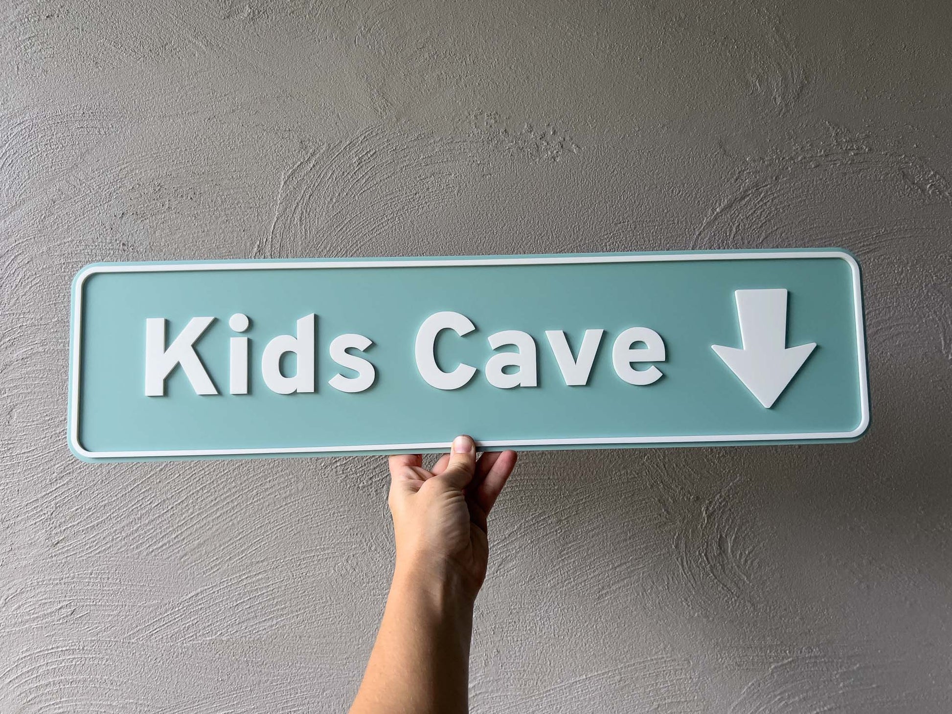 Hand holding a sign that reads 'Kids Cave' with an arrow on a gray wall | Coji Designs