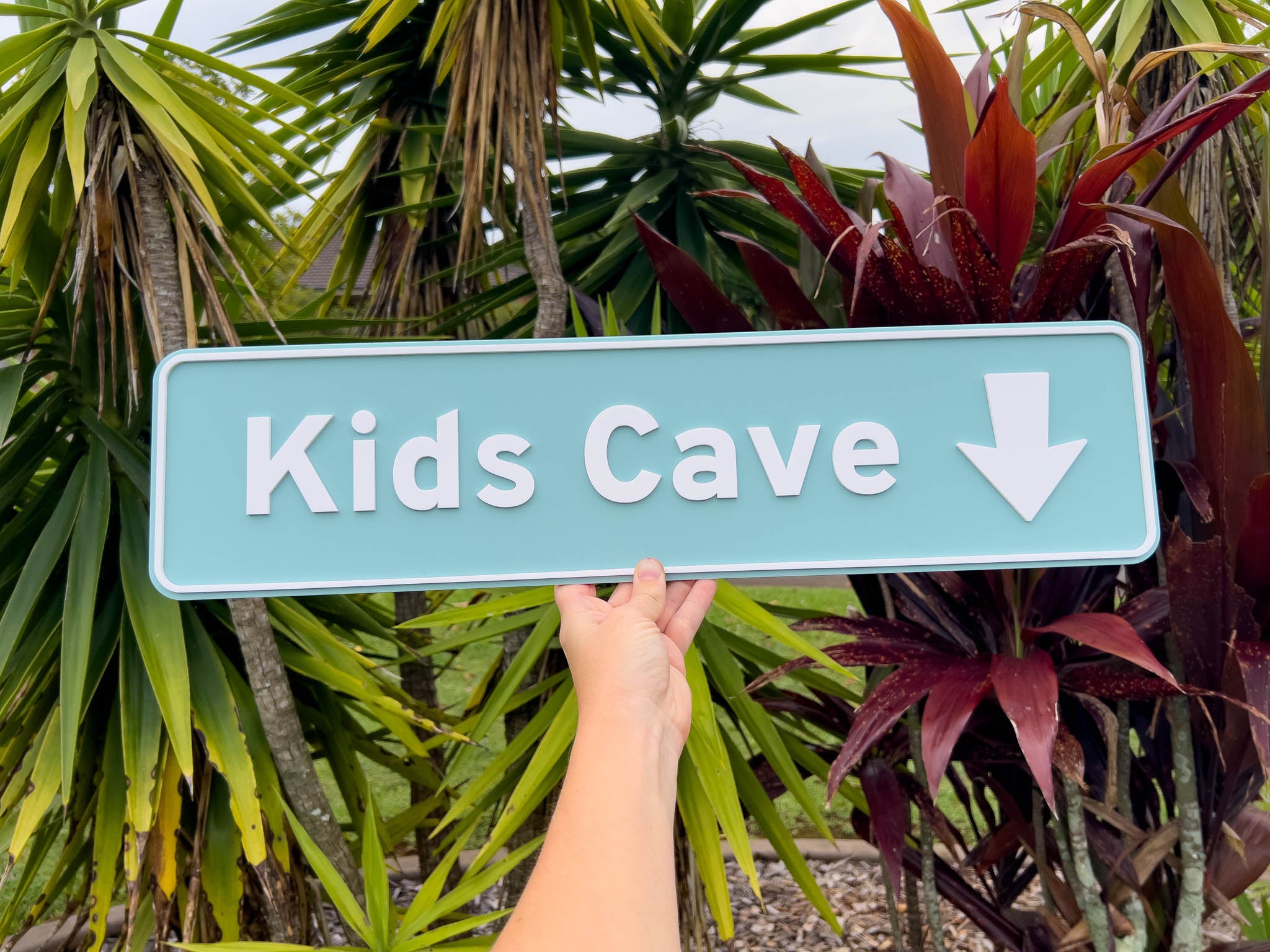 Hand holding a sign that reads 'Kids Cave' with an arrow in front of tropical plants | Coji Designs