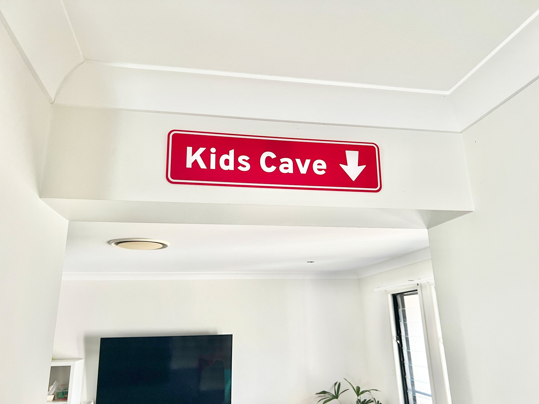 Novelty highway direction sign pointing down into a room that says 'Kids Cave' in a bright ruby red and white colour.
