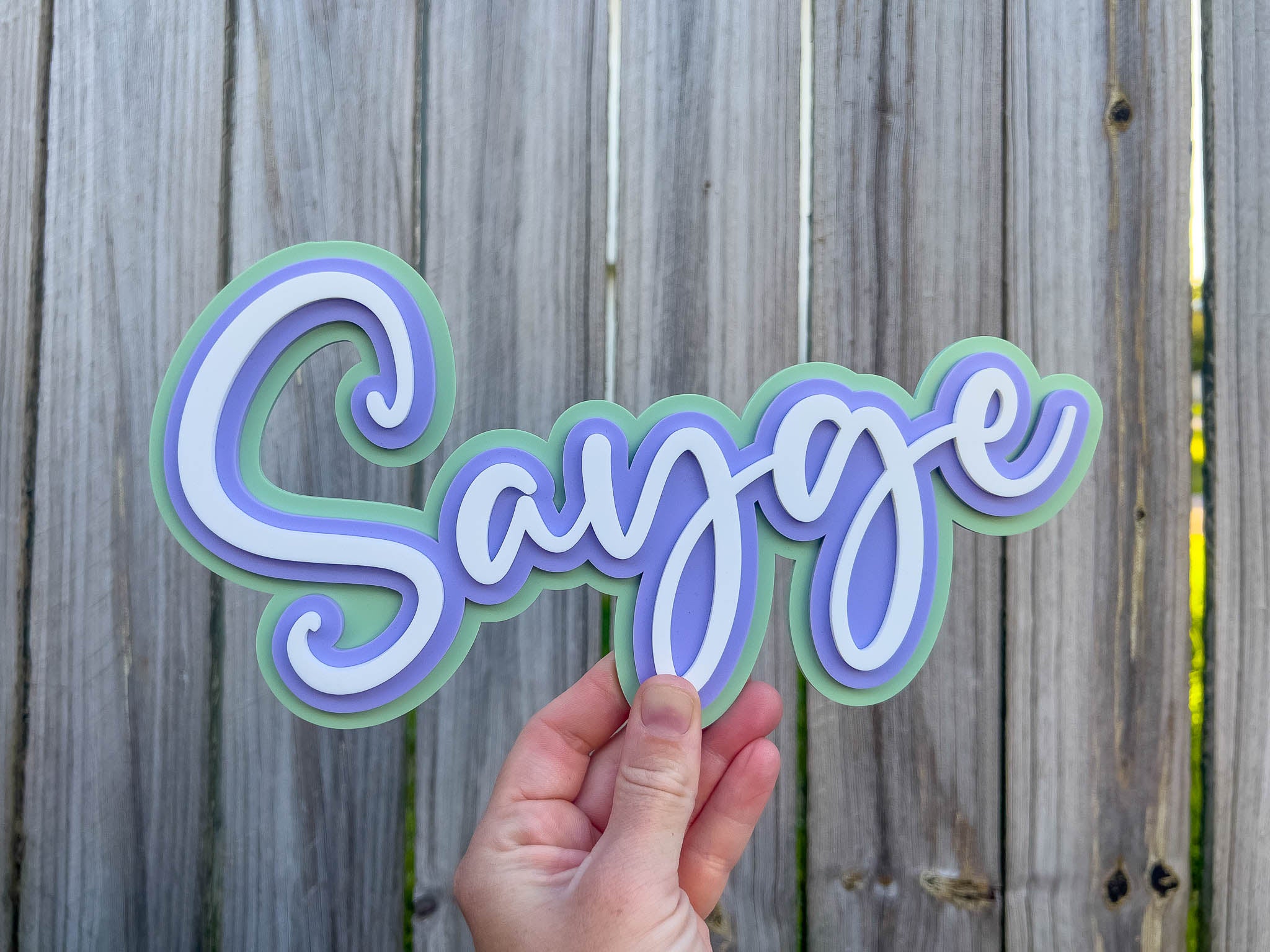 Hand holding a custom cut 3D acrylic name sign with 'Sayge' in wavy letters against a wooden fence background