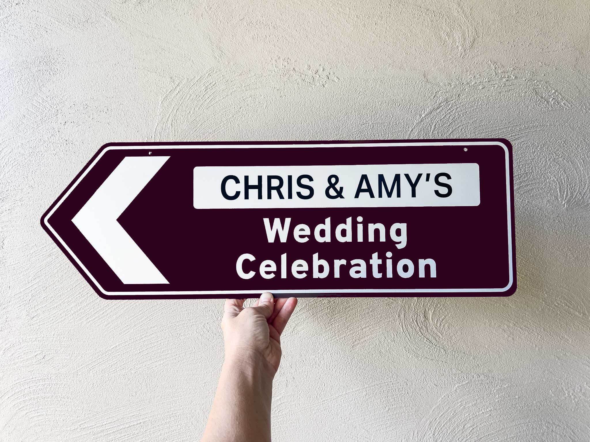 Hand holding a highway style sign with 'Chris & Amy's Wedding Celebration' text against a light wall.
