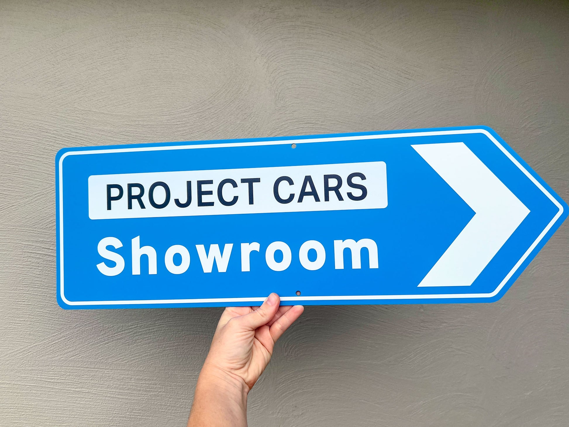 Hand holding a blue sign with 'Project Cars Showroom' text against a gray background