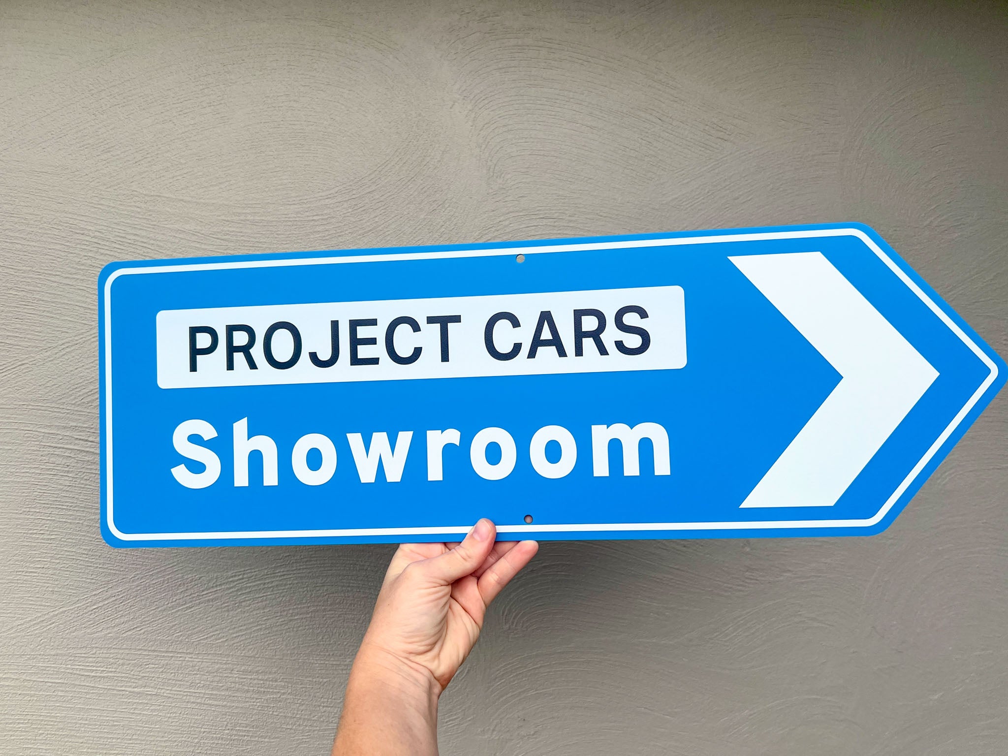 Hand holding a blue sign with 'Project Cars Showroom' text against a gray background
