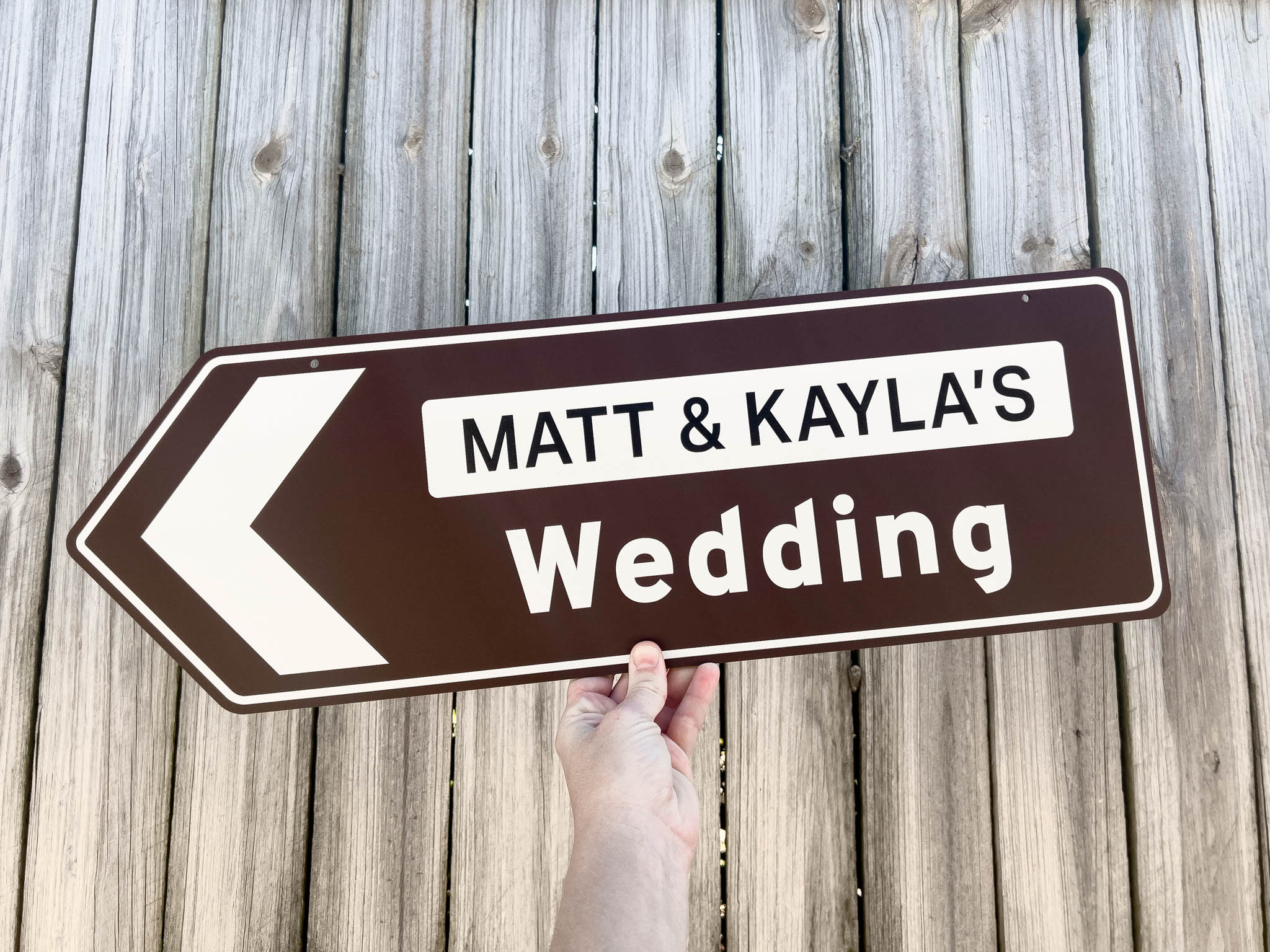 Hand holding a wedding highway direction sign with 'Matt & Kayla's Wedding' text in chocolate brown and white.