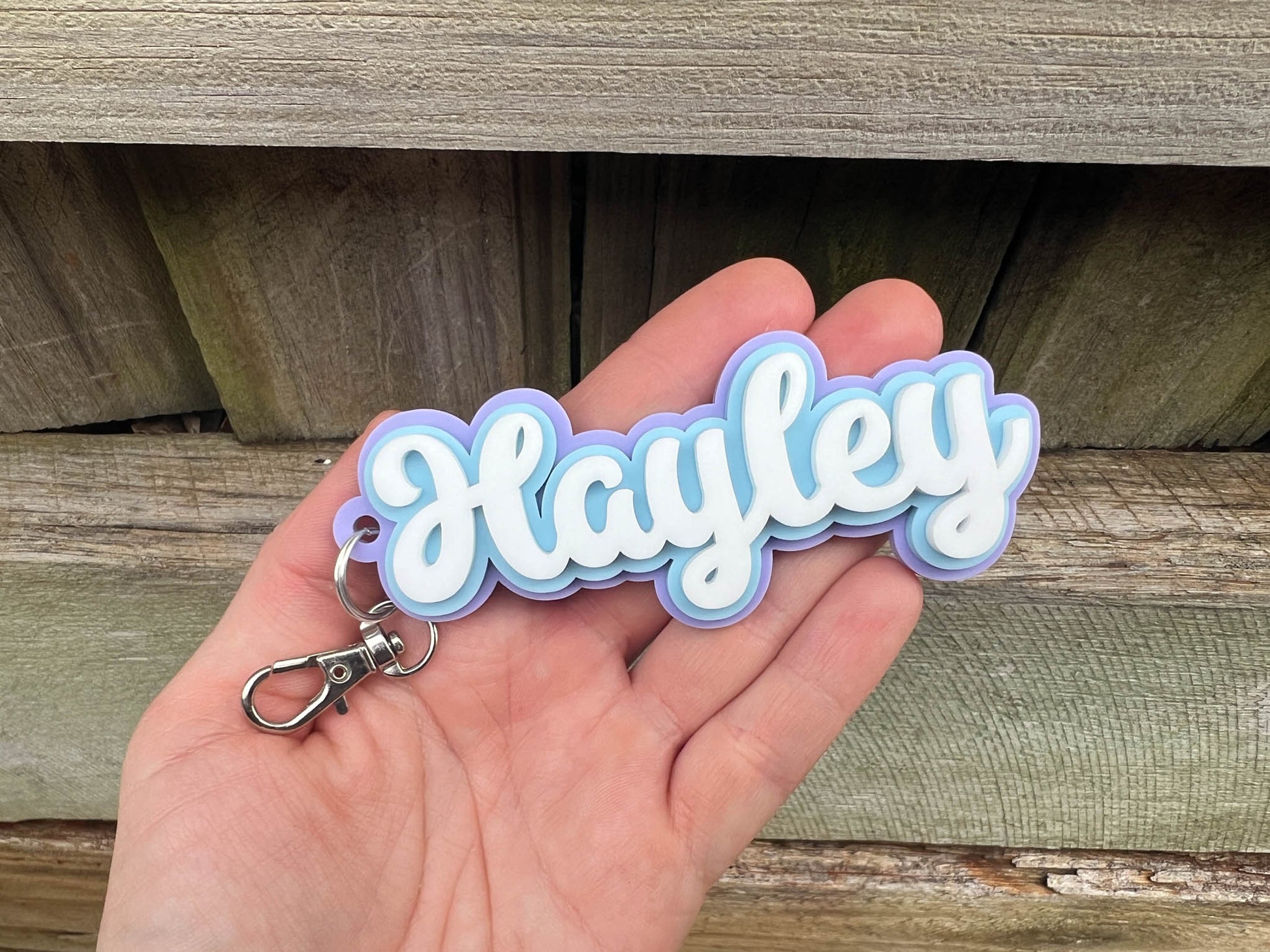 Hand holding a blue, purple and white keychain with 'Hayley' on it against a wooden background