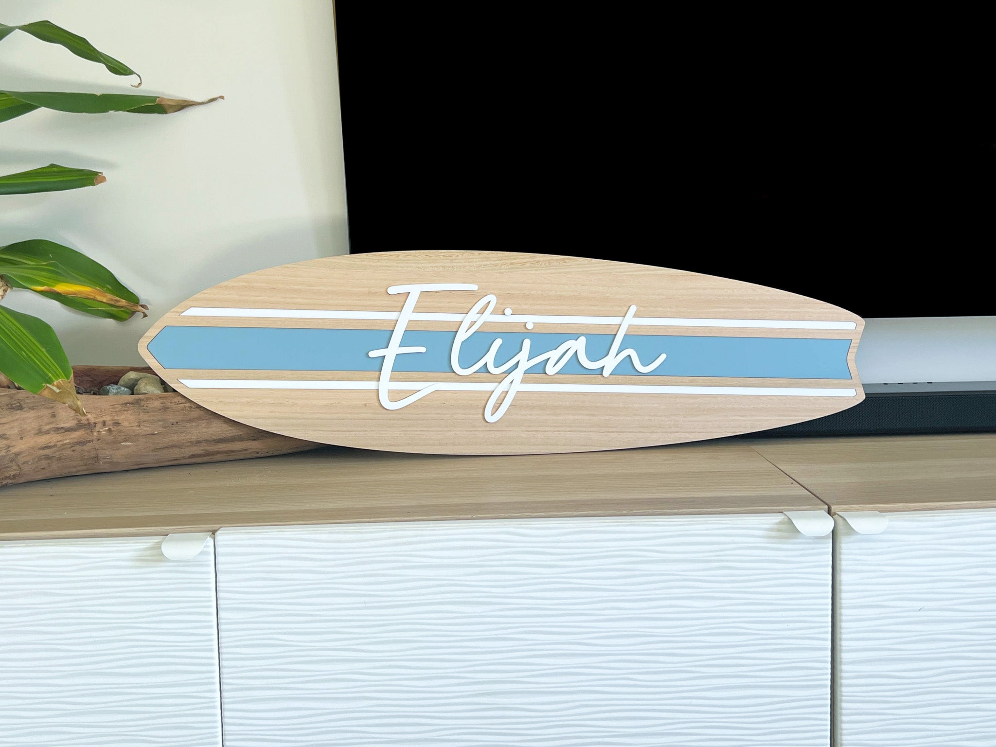Personalised wooden surfboard with a coastal blue stripe and a white 3D name that says 'Elijah' in the centre. 
