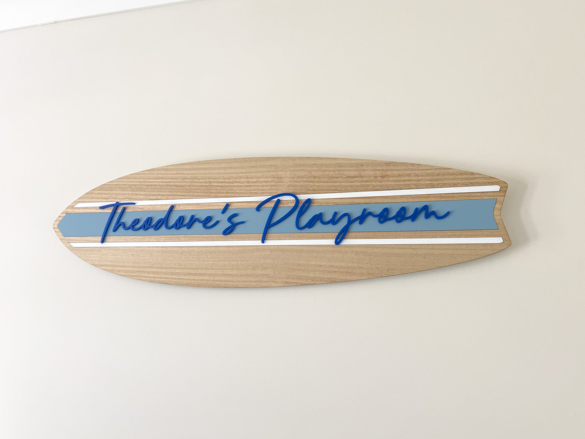 A timber wooden surfboard shaped sign with a dusty blue stripe and deep coastal blue 3D text that says 'Theodore's Playroom hanging on a white wall.