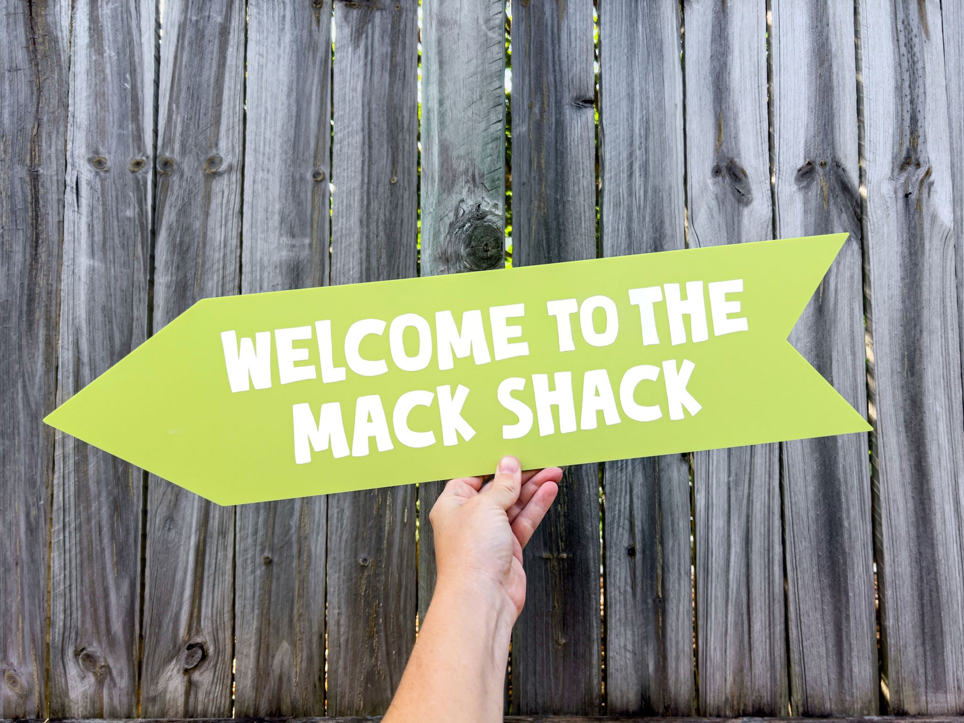 Hand holding a green arrow-shaped sign with 'Welcome to the Mack Shack' text against a wooden fence background.