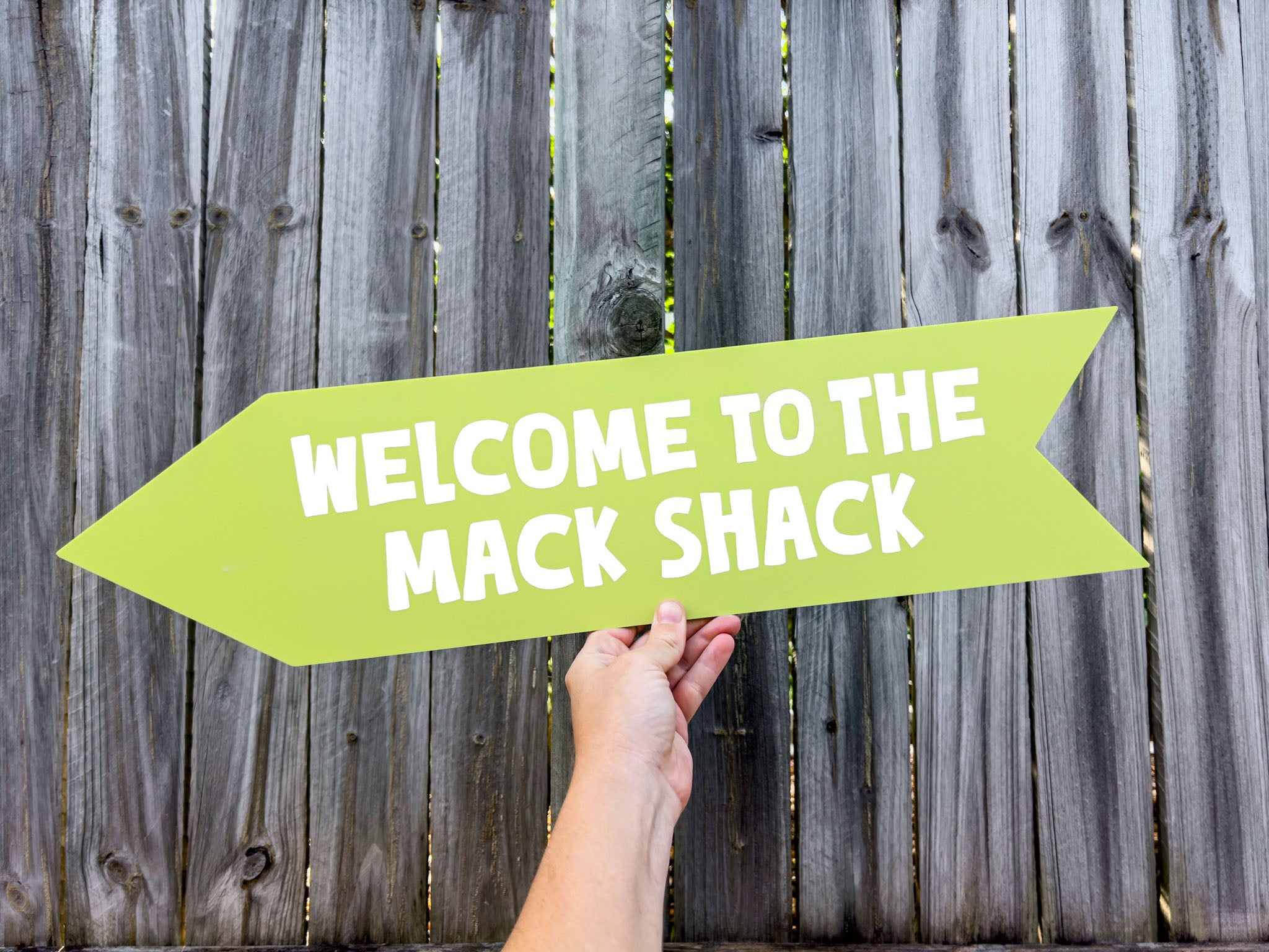 Hand holding a green arrow-shaped sign with 'Welcome to the Mack Shack' text against a wooden fence background.
