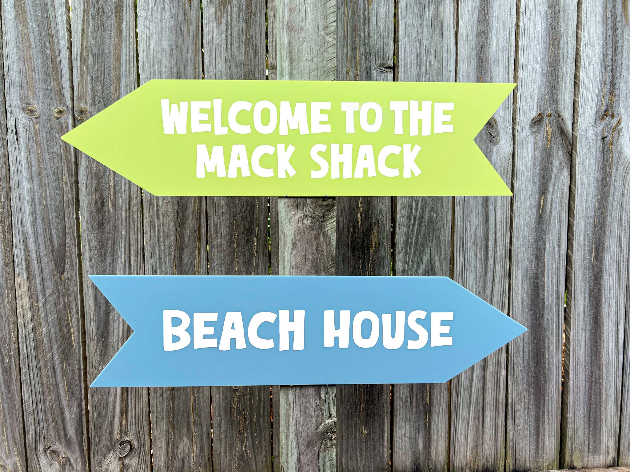 Two directional signs on a wooden background, one green and one blue, with text 'Welcome to the Mack Shack' and 'Beach House'.