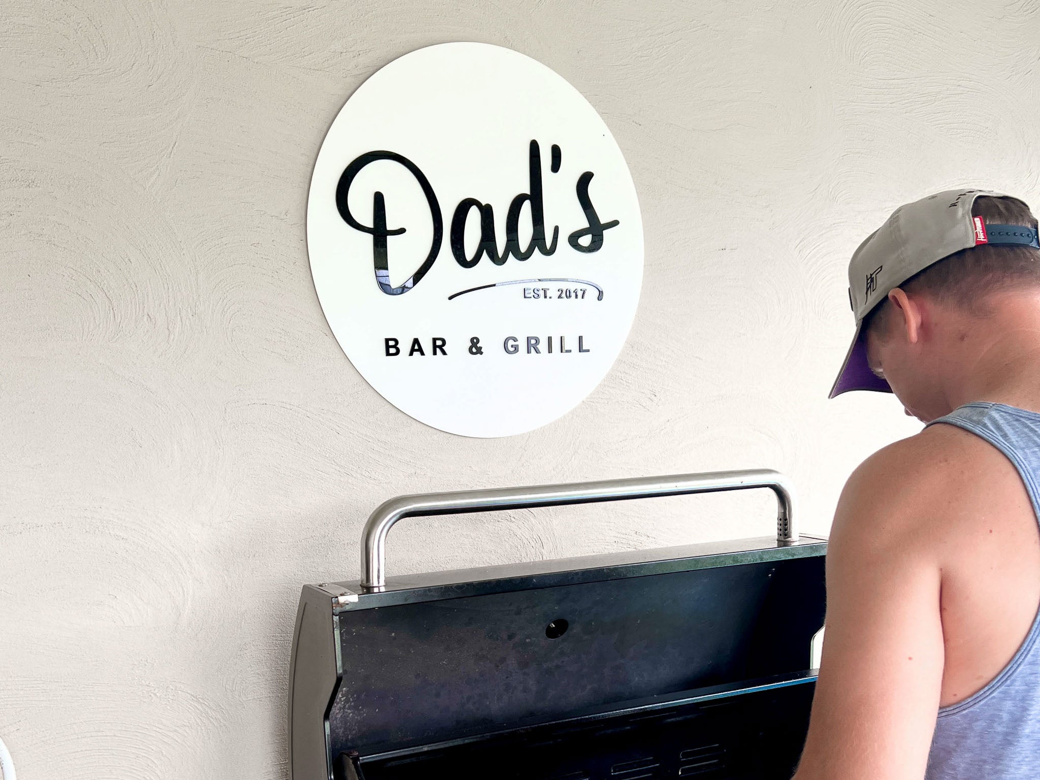 Person grilling on a BBQ with a white and black 'Dad's Bar & Grill' sign on wall