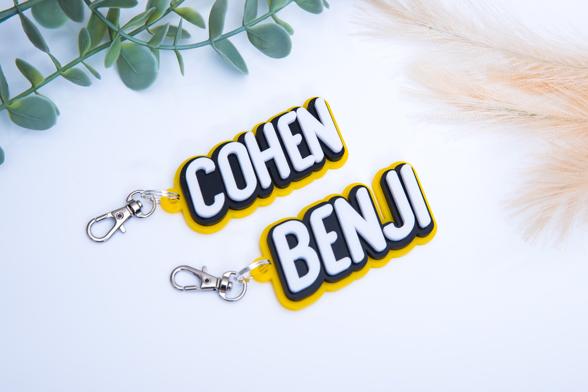 Two bold font 3D acrylic keychains in yellow, black and white that read 'COHEN' & 'BENJI'.