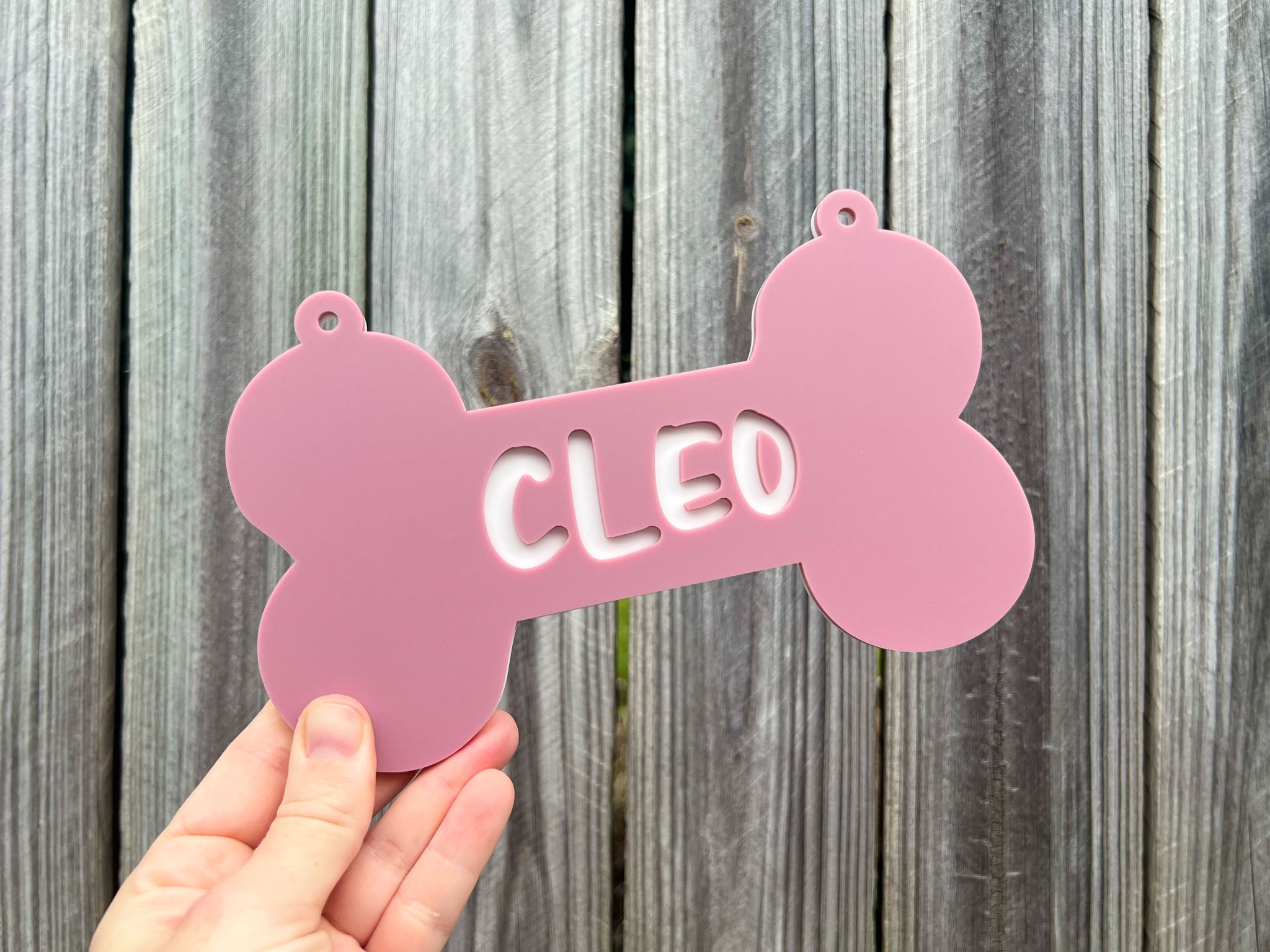 Pink bone-shaped pet tag with 'CLEO' on it held against a wooden background