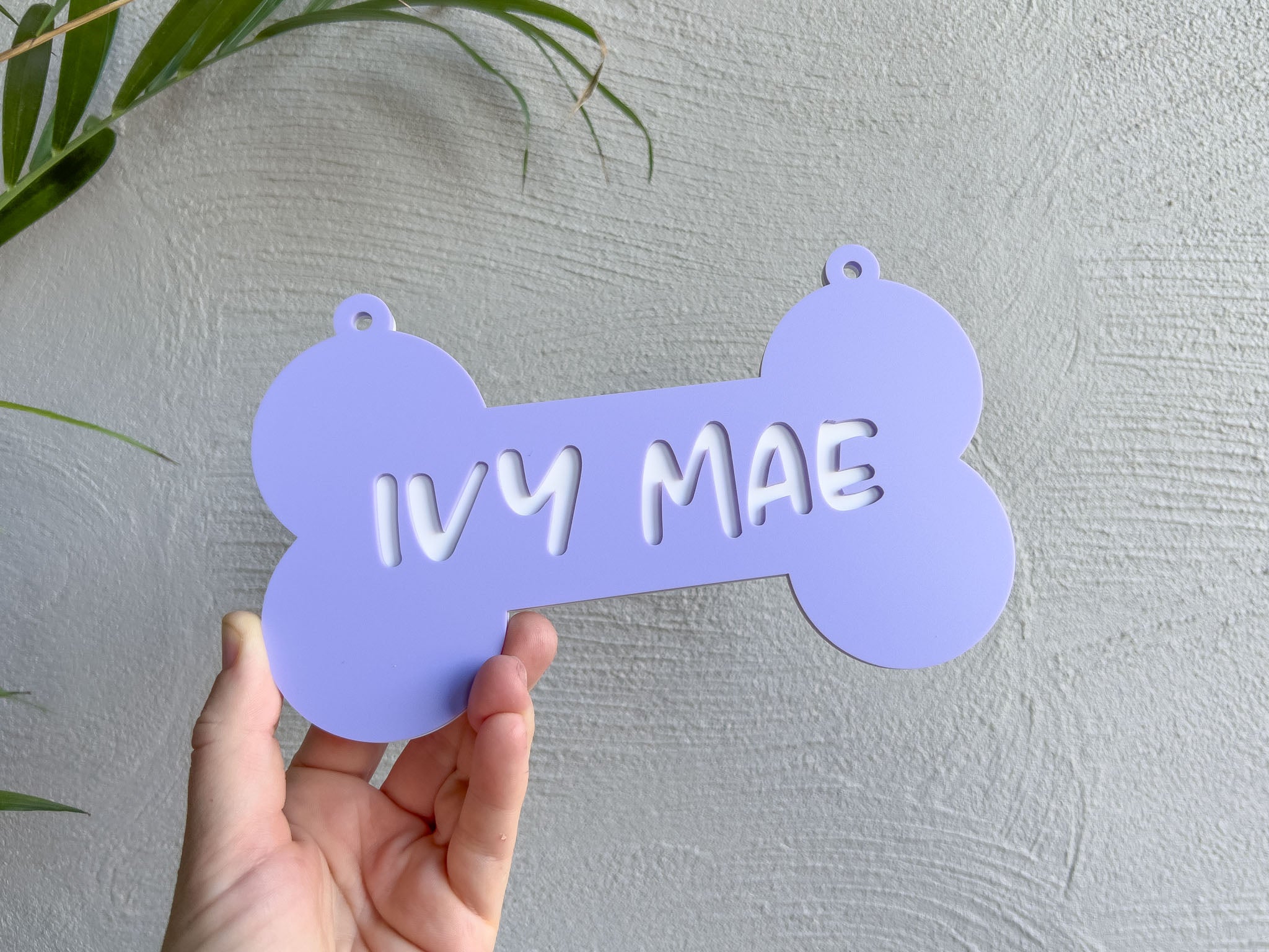 Hand holding a purple bone-shaped sign with 'IVY MAE' text against a light gray background | Coji Designs