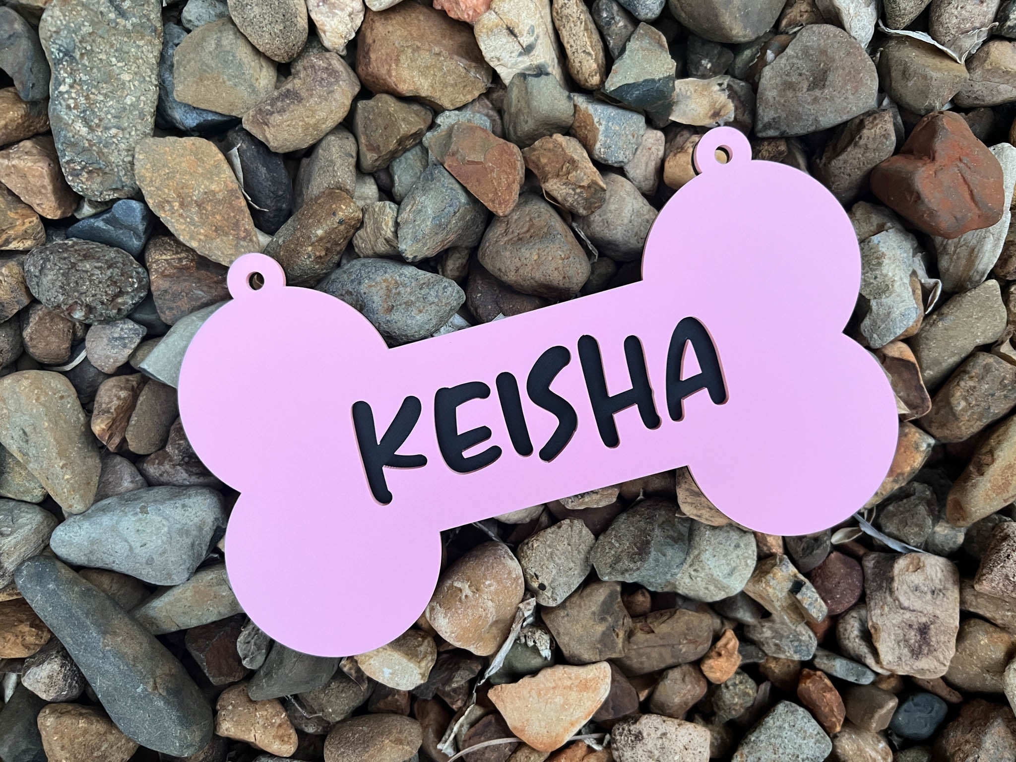 Pink bone-shaped pet tag with 'KEISHA' on it, placed on a bed of small stones | Coji Designs