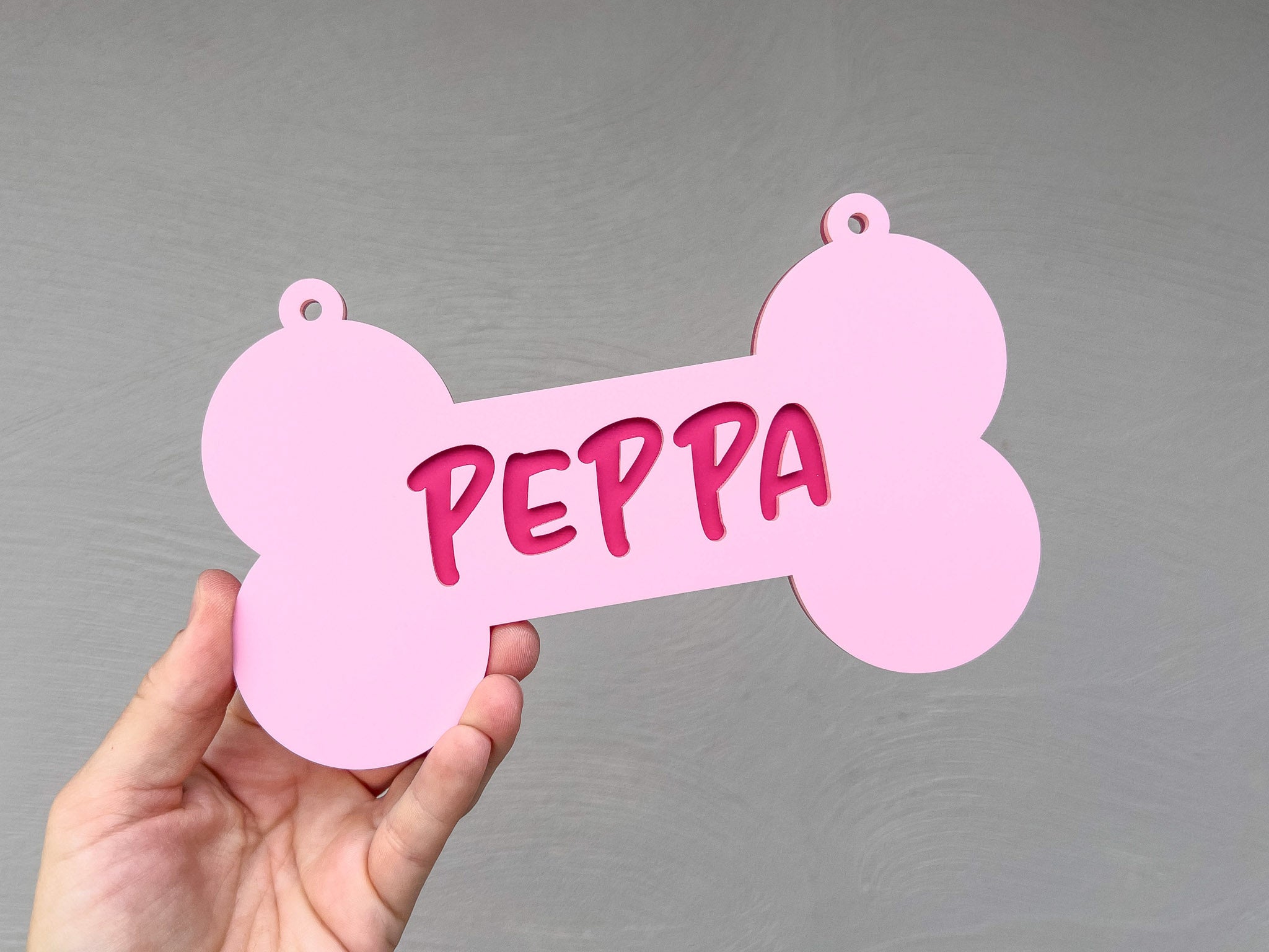 Hand holding a pink bone-shaped tag with 'PEPPA' written on it against a gray background