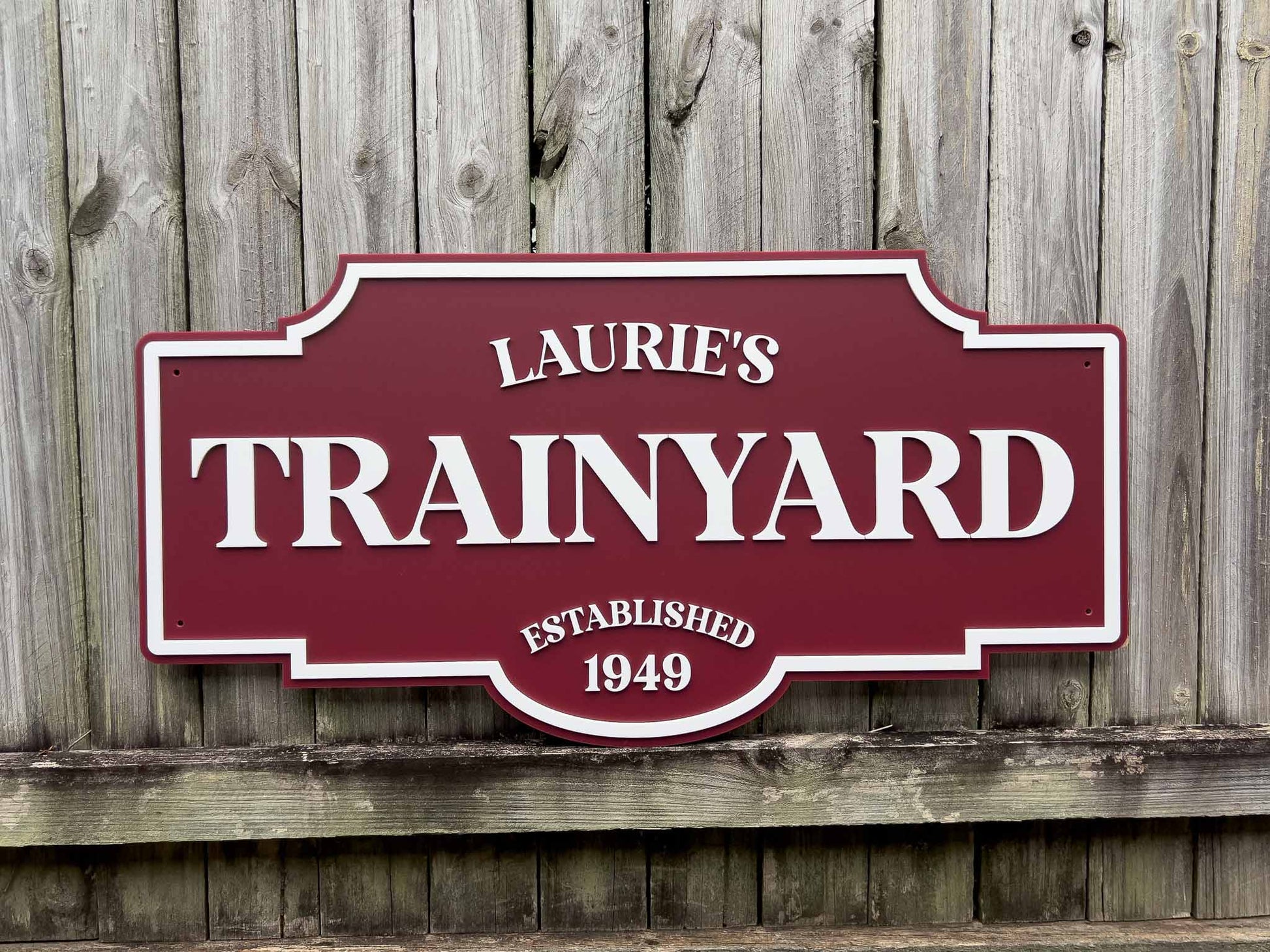 Handcrafted acrylic maroon and white sign with 'Laurie's Trainyard' on a wooden background.