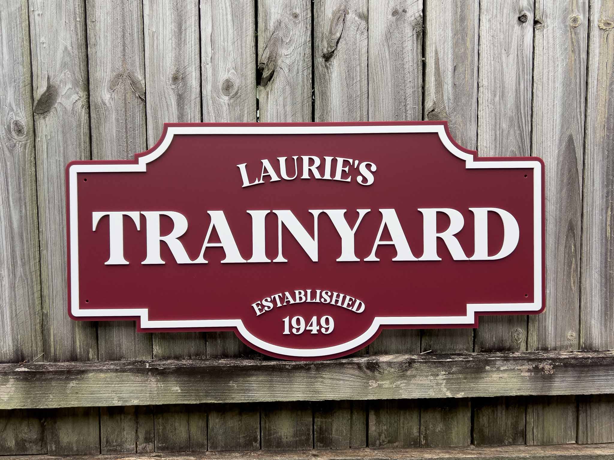 Handcrafted acrylic maroon and white sign with 'Laurie's Trainyard' on a wooden background.