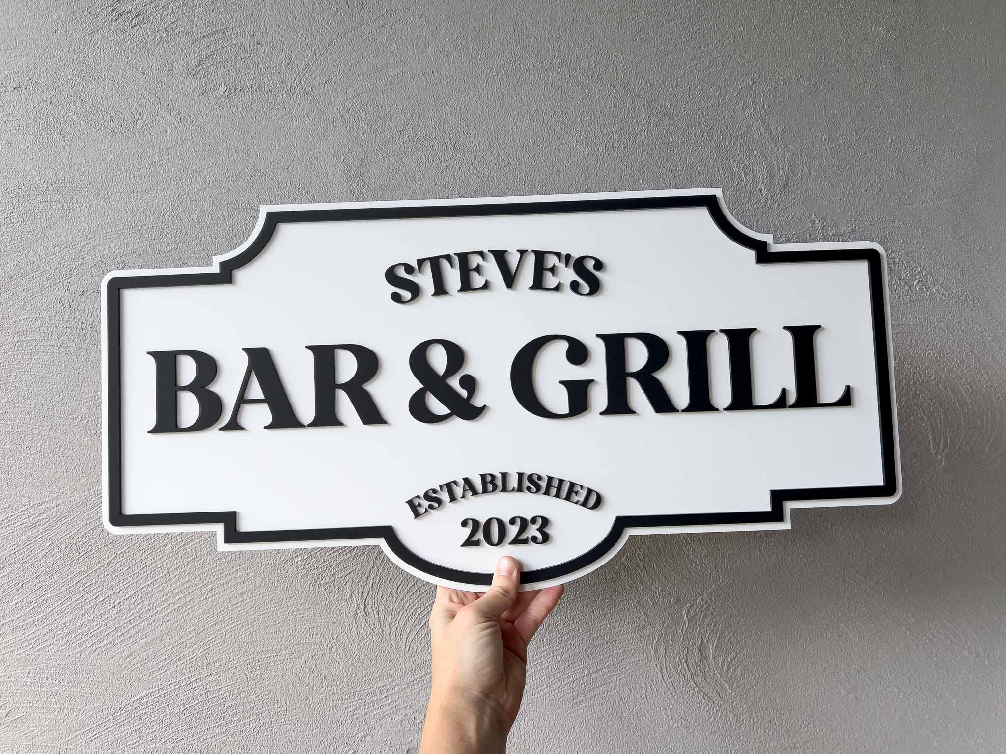 Hand holding a vintage style sign that reads 'Steve's Bar & Grill Established 2023' against a gray background