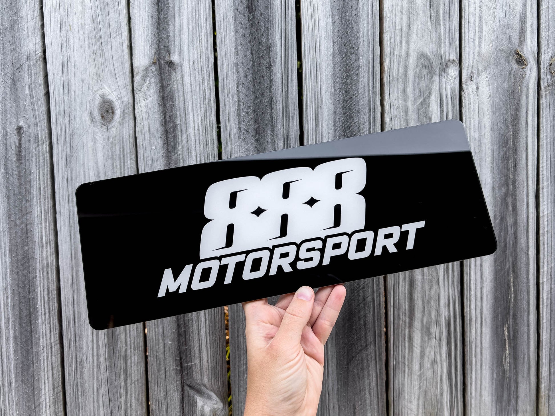 Hand holding a black and white logo plate with '888 Motorsport' logo against a wooden background