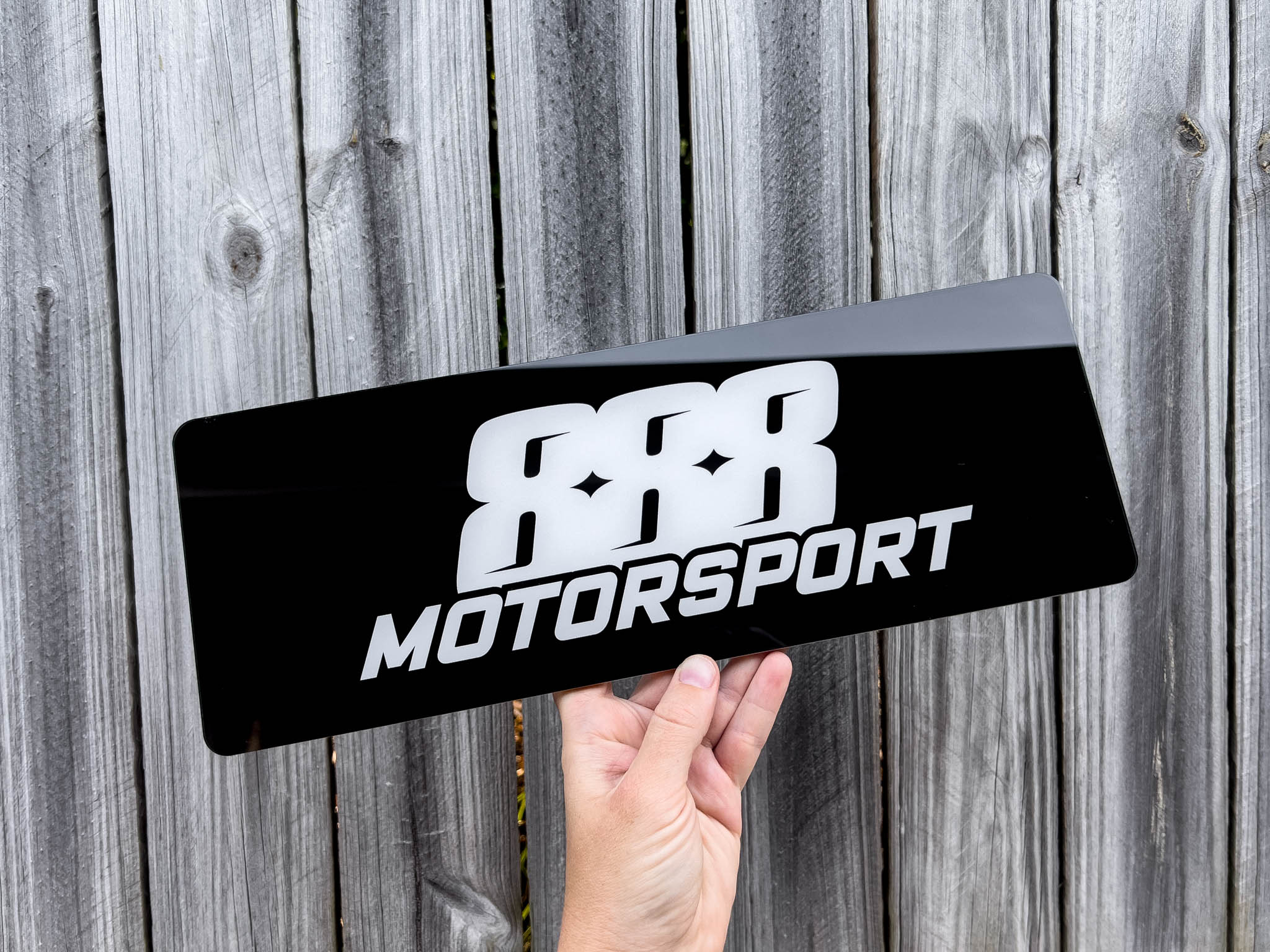 Hand holding a black and white logo plate with '888 Motorsport' logo against a wooden background