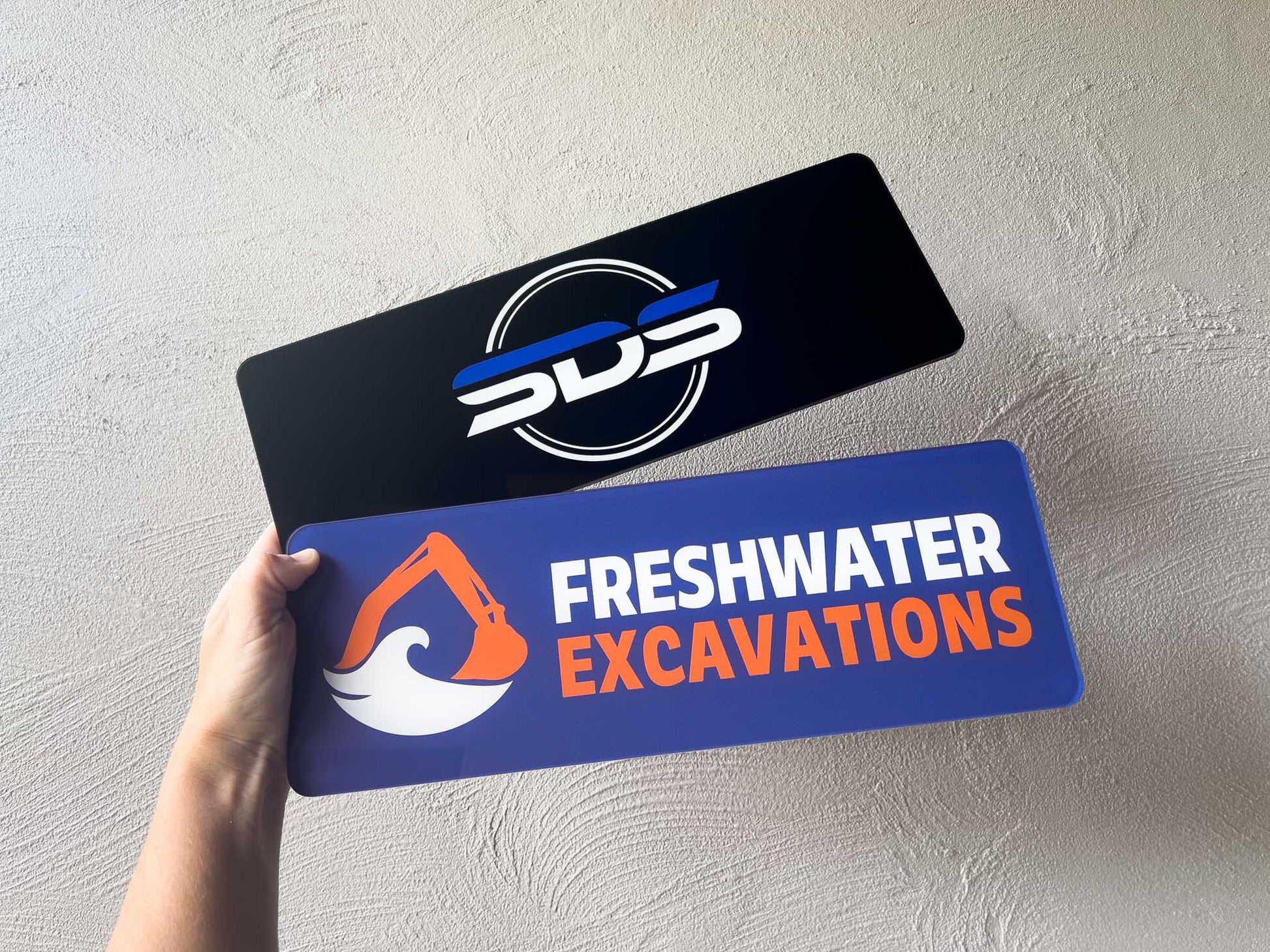Two signs with logos and text, one black with a white and blue logo, the other blue with orange and white text, held against a light gray background.
