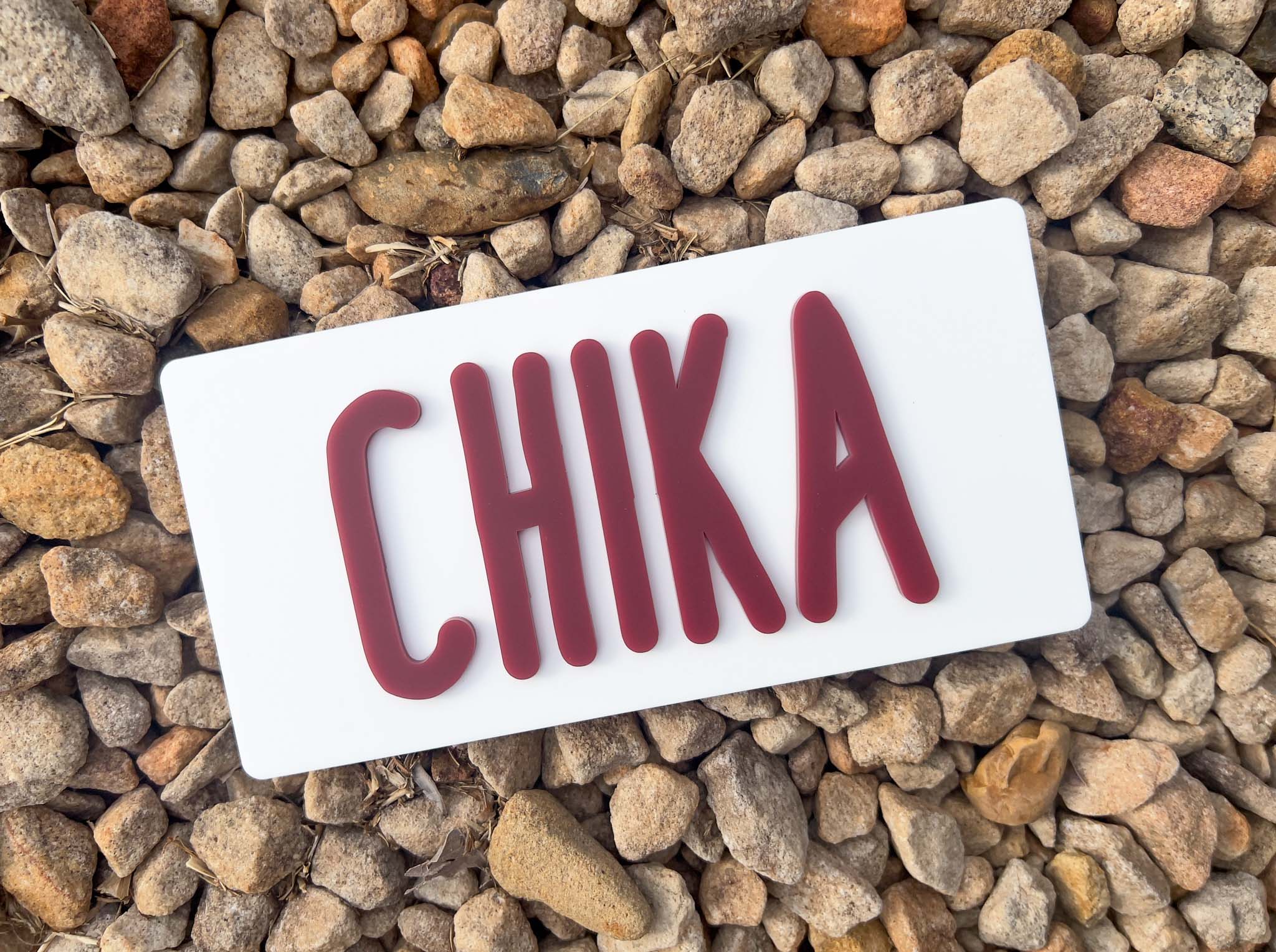 Rectangular white sign with 'CHIKA' in red letters on a background of small stones | Coji Designs